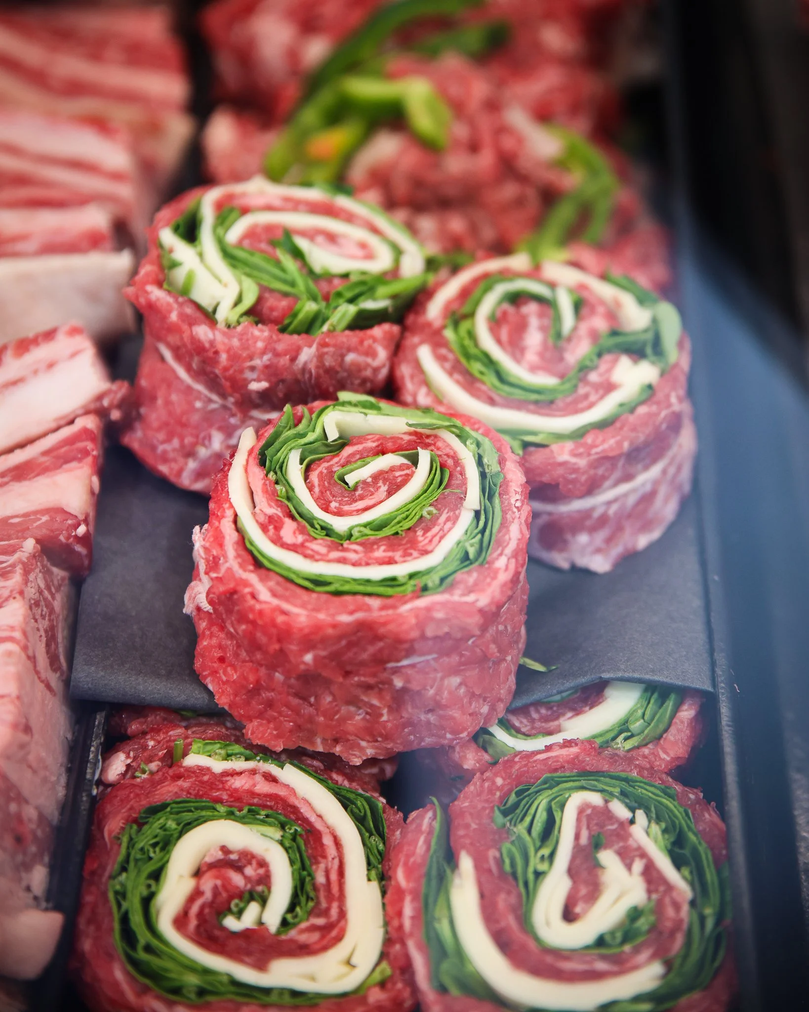 Raw rolled meat with layers of green and white vegetables, arranged in a spiral pattern, on a black surface.