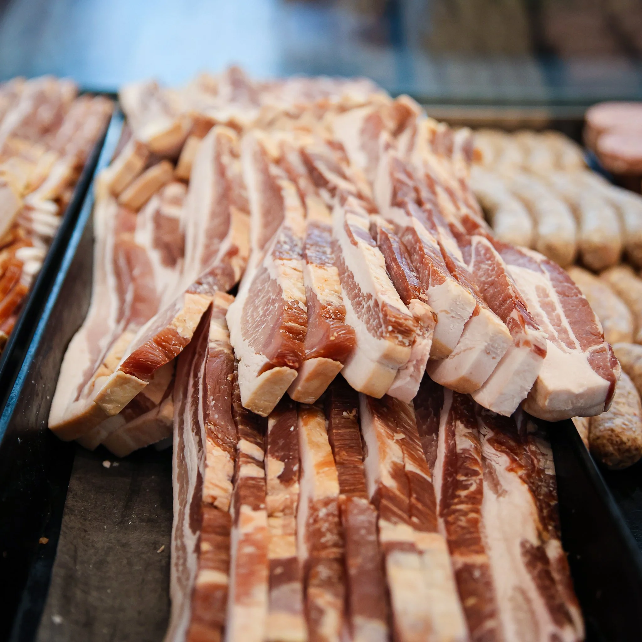 Slices of bacon arranged on a black tray at a butcher shop or deli.