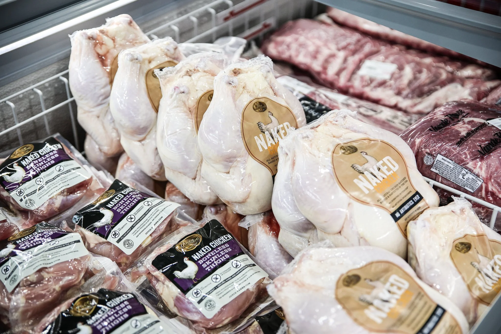 Frozen chicken parts, including whole chickens and packages of chicken thighs displayed in a supermarket freezer.