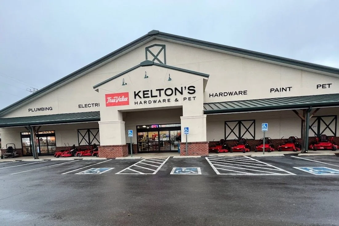 Exterior view of Kelton's Hardware and Pet store with a parking lot in front, including accessible parking spaces and several red riding lawn mowers lined up under the covered area