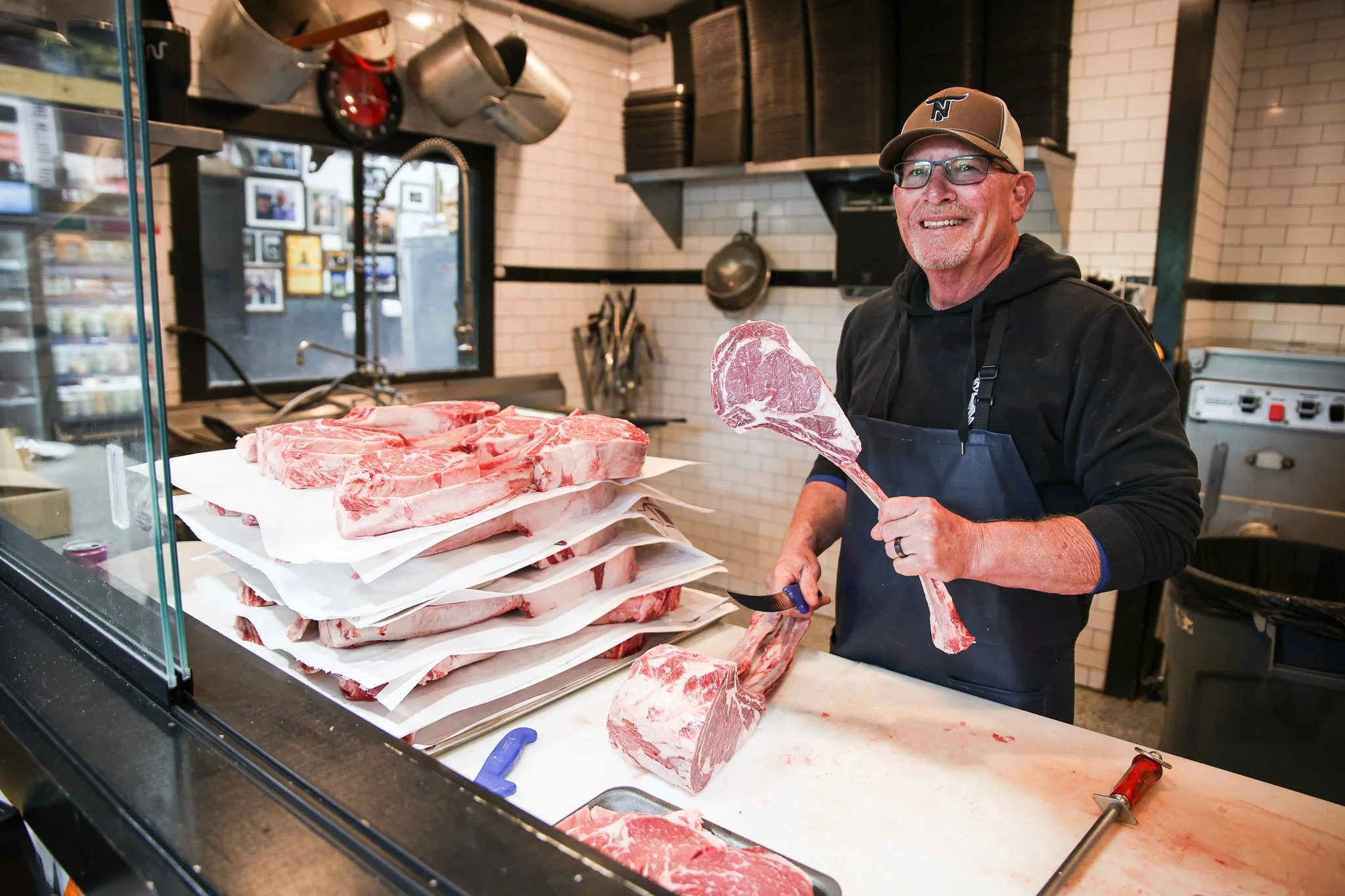 A smiling man wearing glasses, a baseball cap, and a black hoodie stands behind a butcher counter with raw meat, holding a large piece of meat on a cleaver. There are multiple stacks of raw meat on the counter and various kitchen utensils and pots ha
