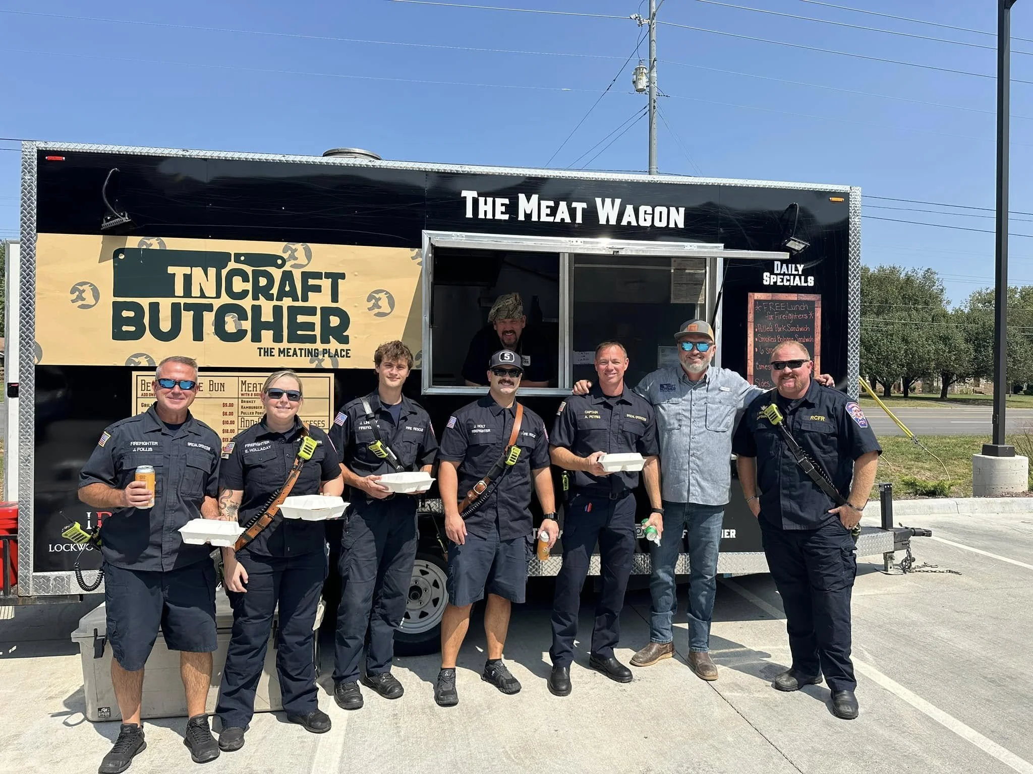 Group of seven people, including police officers and civilians, standing in front of a food truck called "The Meat Wagon" and "TN Craft Butcher." Three police officers in uniform are holding food containers, while two civilians, one in sunglasses and