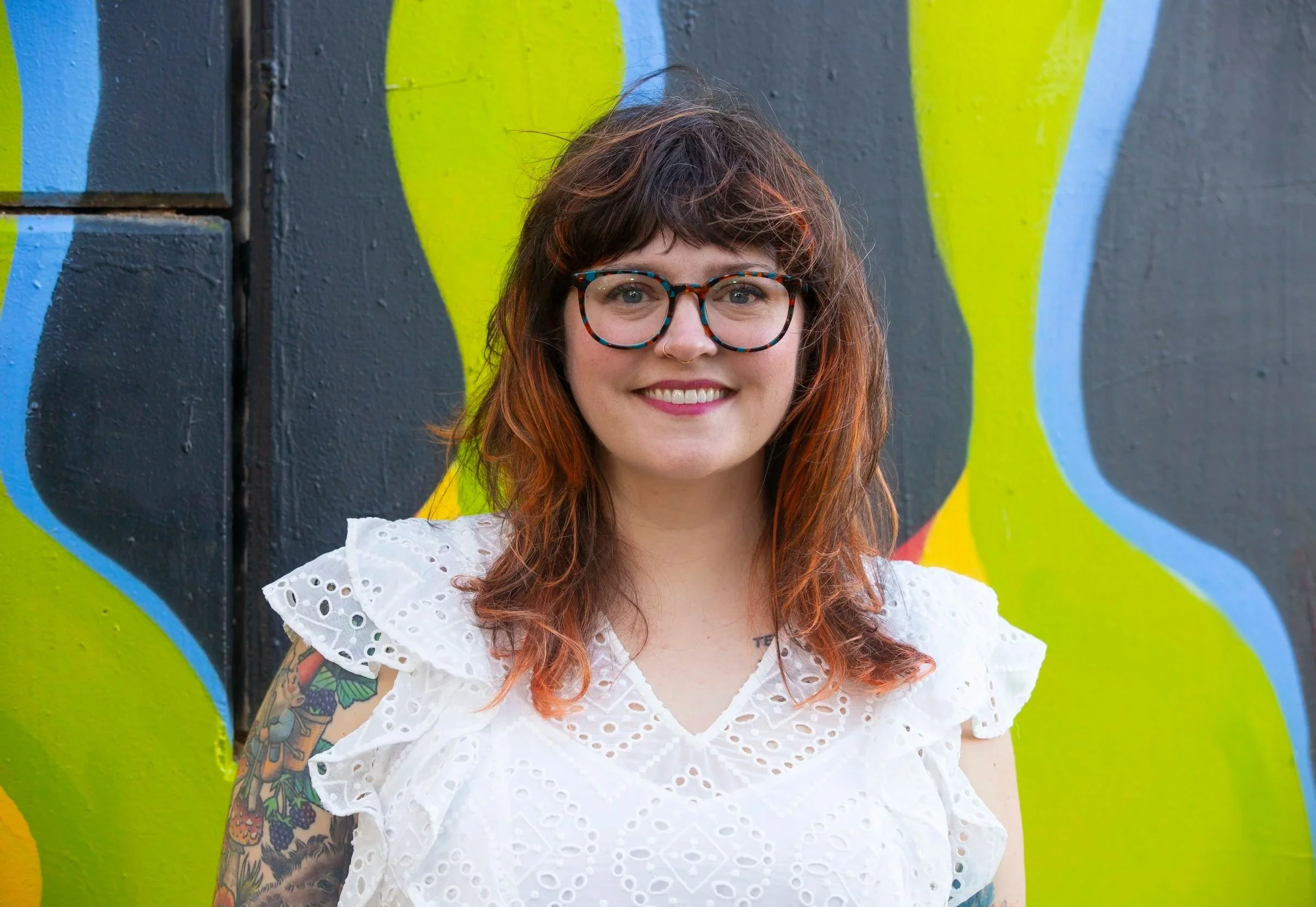 Headshot of Trina Bolfing - a trauma-informed therapist in Austin, Texas - with glasses and curly reddish hair standing in front of a colorful graffiti mural.