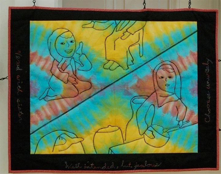 Colorful tie-dye quilt with embroidered outlines of two women, one holding a bowl and the other sitting with a crossbody bag, and two peace signs, with handwritten text on the black border.