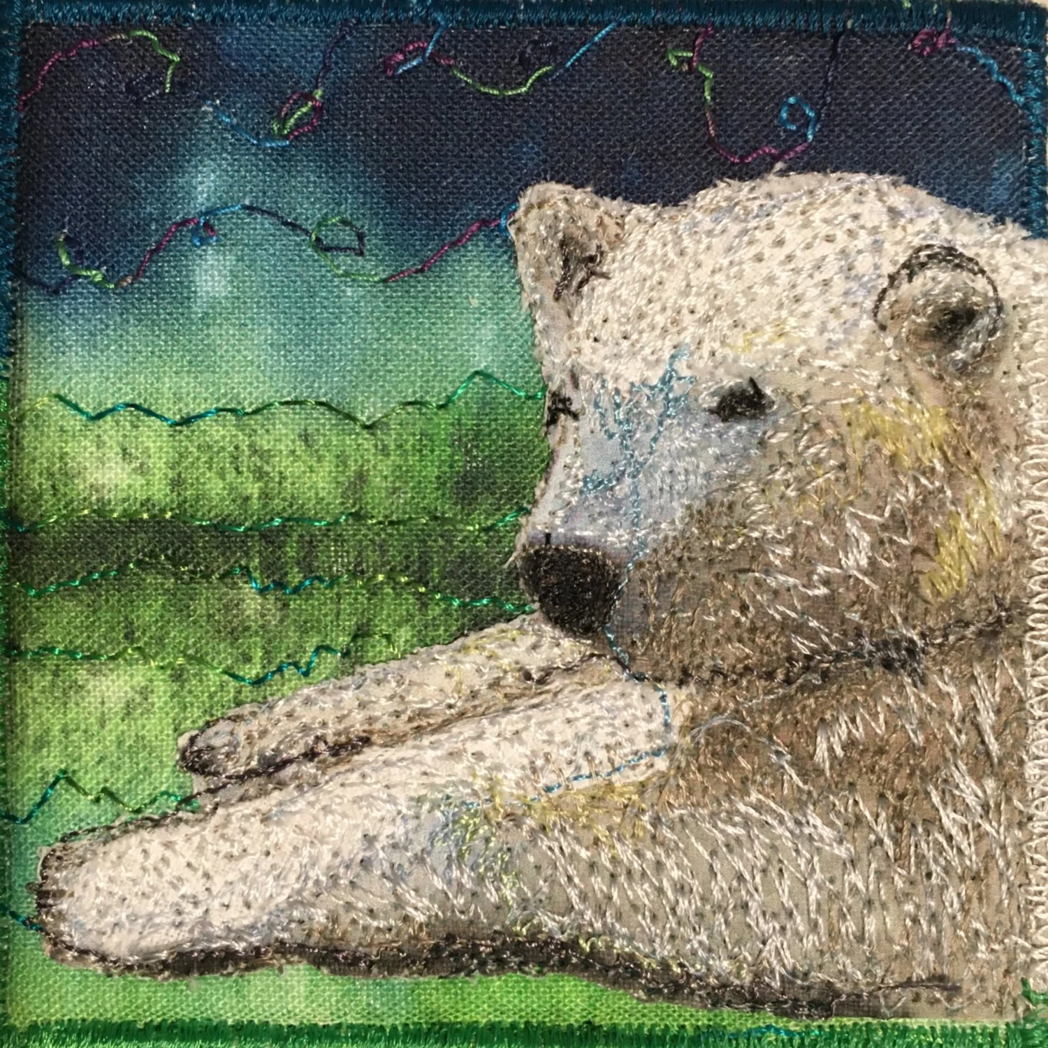 Quilted artwork of a white polar bear resting on a green landscape with a blue sky.