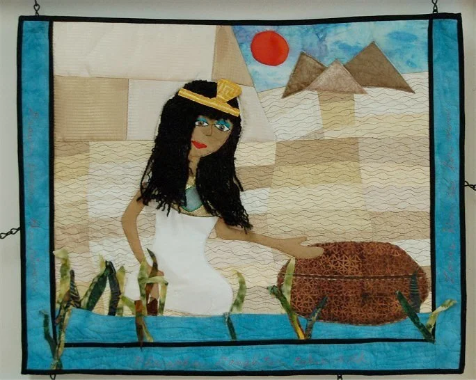 A quilted wall hanging depicting an Egyptian scene with a woman in a white dress, black hair, a yellow headband, and a turquoise necklace, standing near the Nile River with pyramids and the sun in the background.