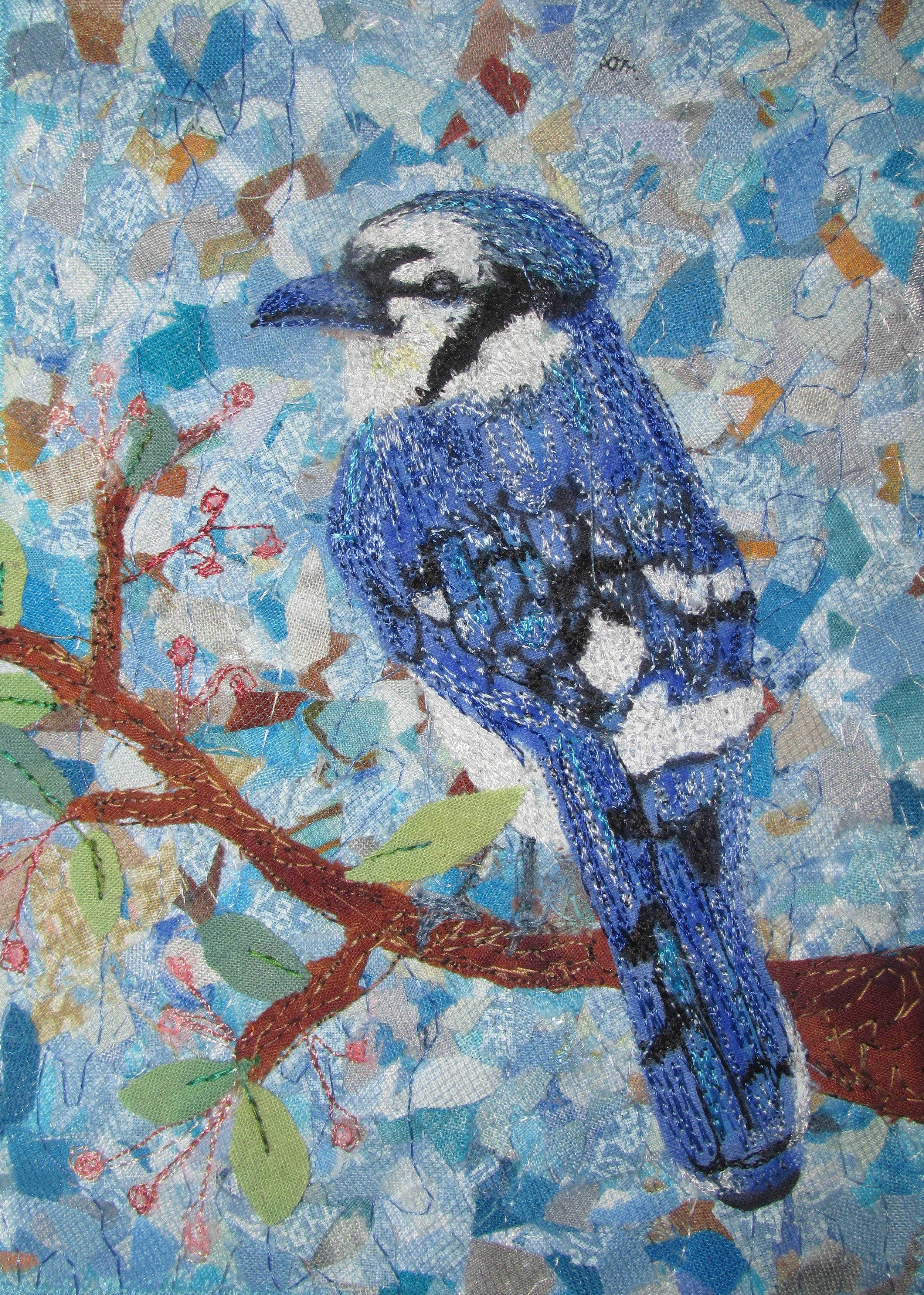 Art quilt of a abird perched on a branch with green leaves and red berries, set against a background of various shades of blue and beige fabrics.