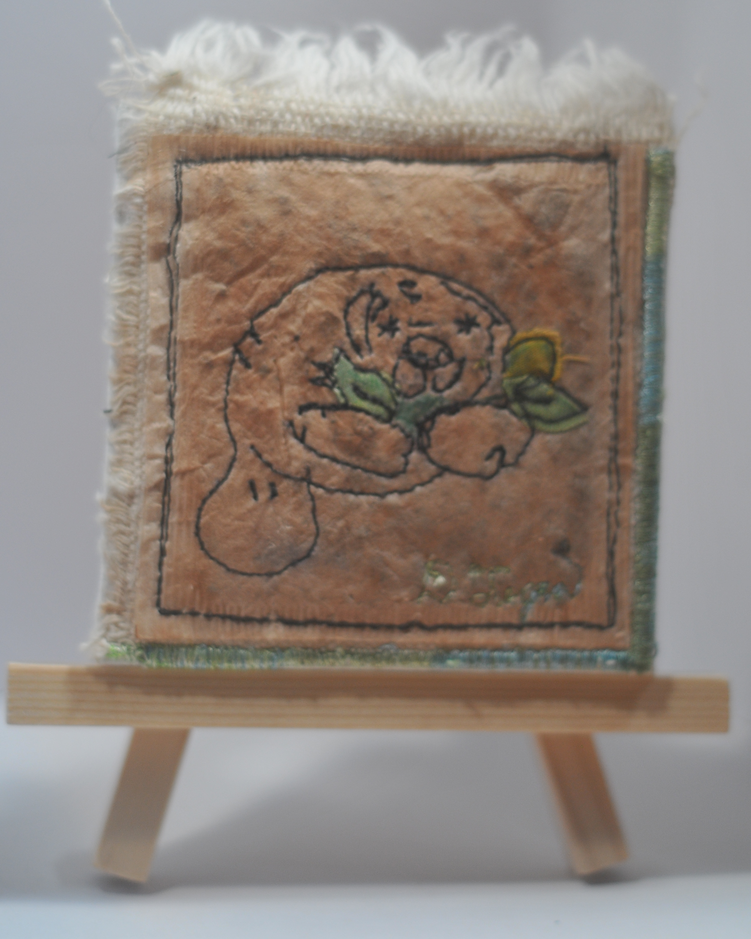 A small fabric quilt with a drawing of a manatee holding a flower, mounted on a miniature wooden easel.