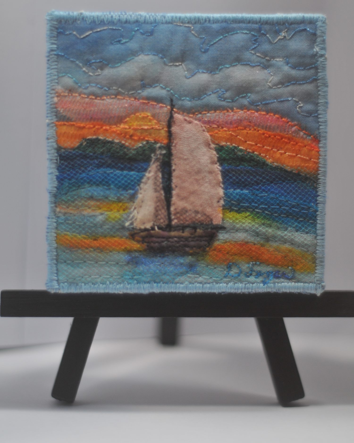 artwork of a sailing boat on water with a sunset and mountains in the background, framed with a blue border, displayed on a small black easel.