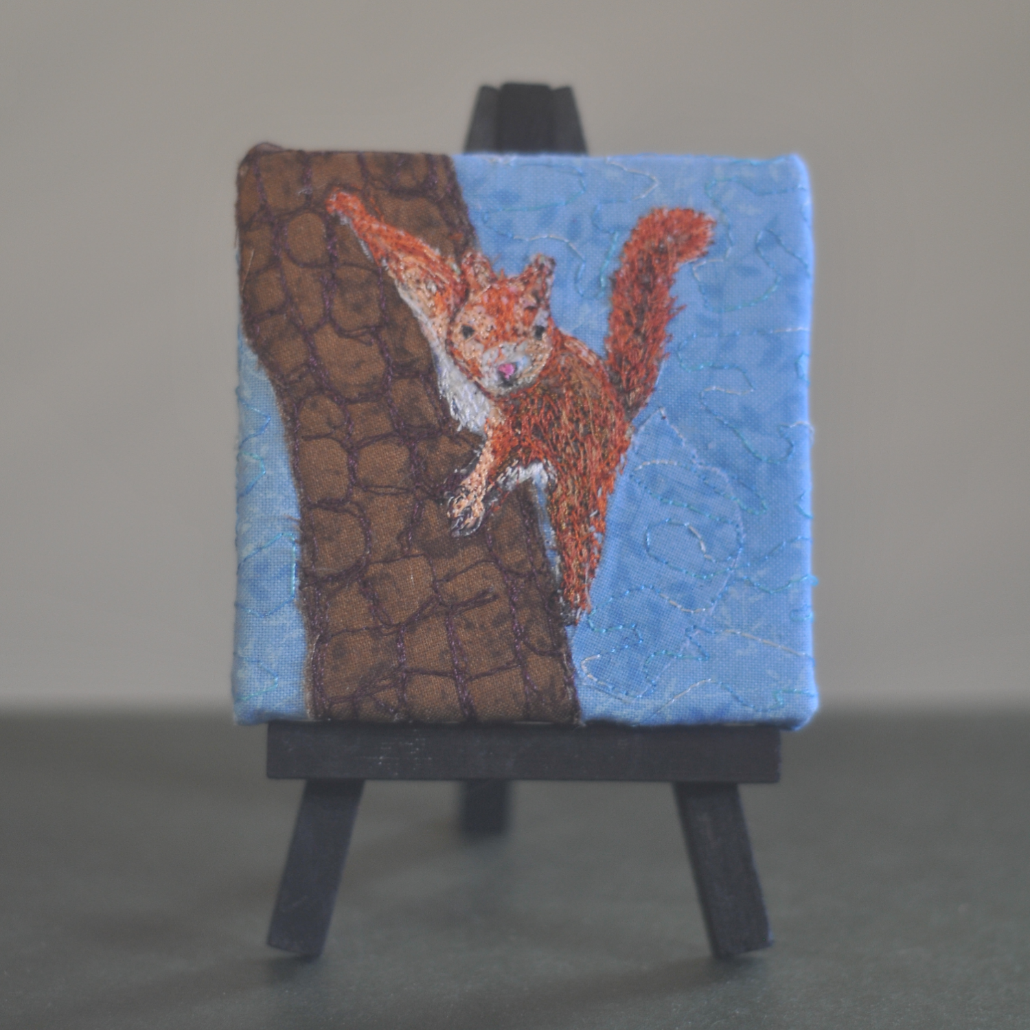 Quilted artwork of a squirrel climbing a tree on a small canvas, displayed on a miniature easel.