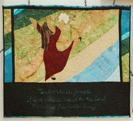 Quilt wall hanging depicting a woman in a red dress walking on a path beside a river, with handwritten text beneath.