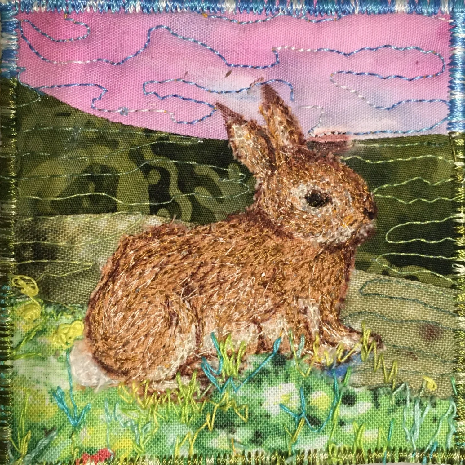 Fabric art bookmarker of a rabbit sitting on grass with pink clouds and blue sky in the background.