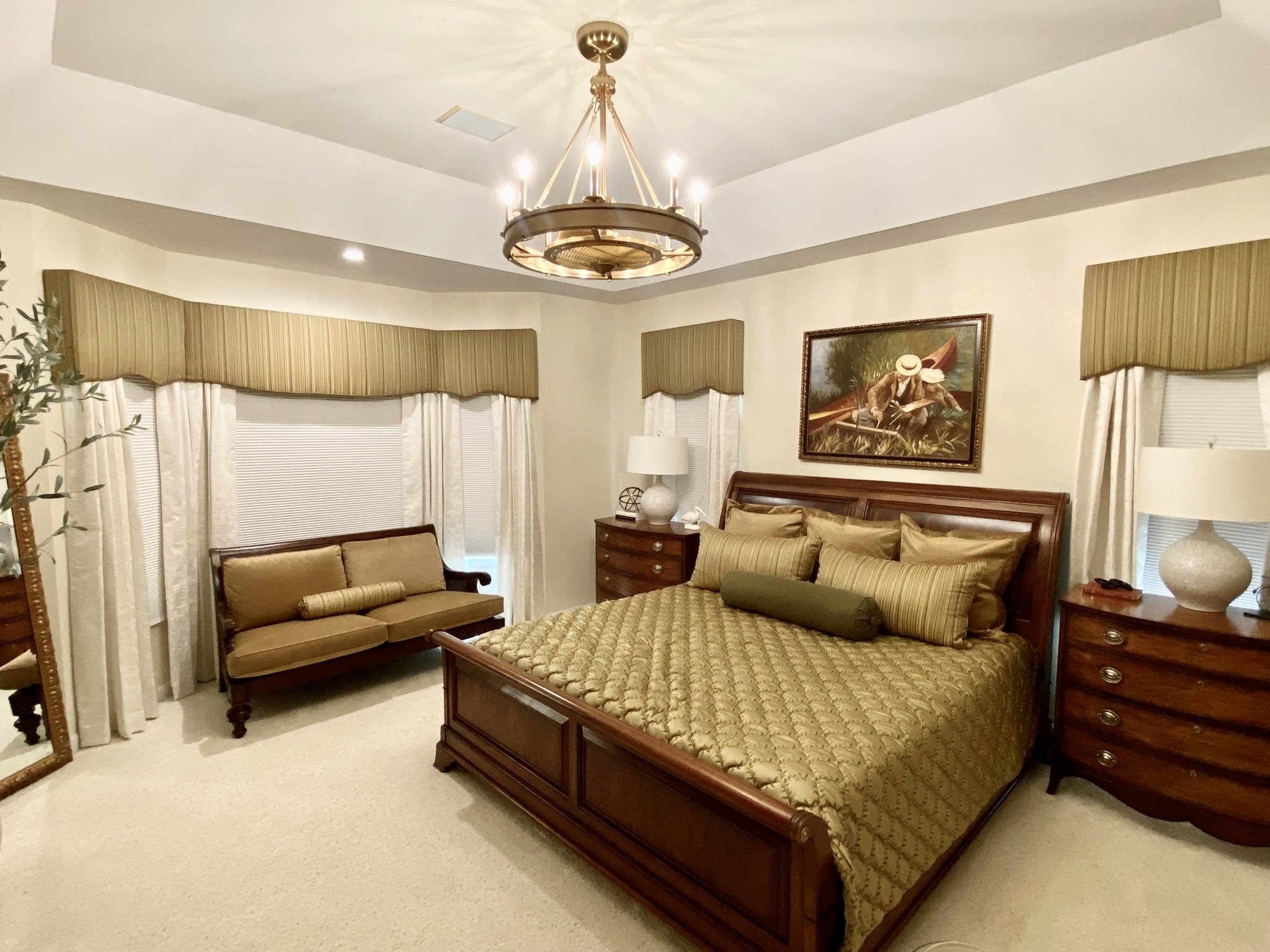 A traditional elegant master primary bedroom with cream and gold custom drapes and bedding, a gold fandelier, with Ethan Allen Furniture pieces. A large gold mirror and pottery barn olive tree in large pot decorates the room with Perigold white lamps