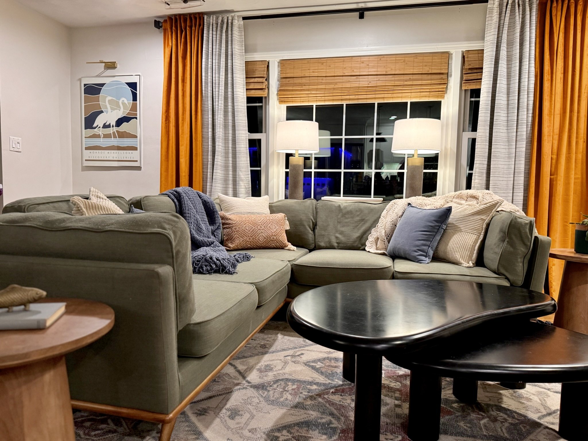 Cozy living room with a green sectional Article sofa, decorated with throw blue throw pillows, beige corduroy throw pillows and blankets. West Elm nesting black coffee table, orange velvet curtains and black and white striped curtains.