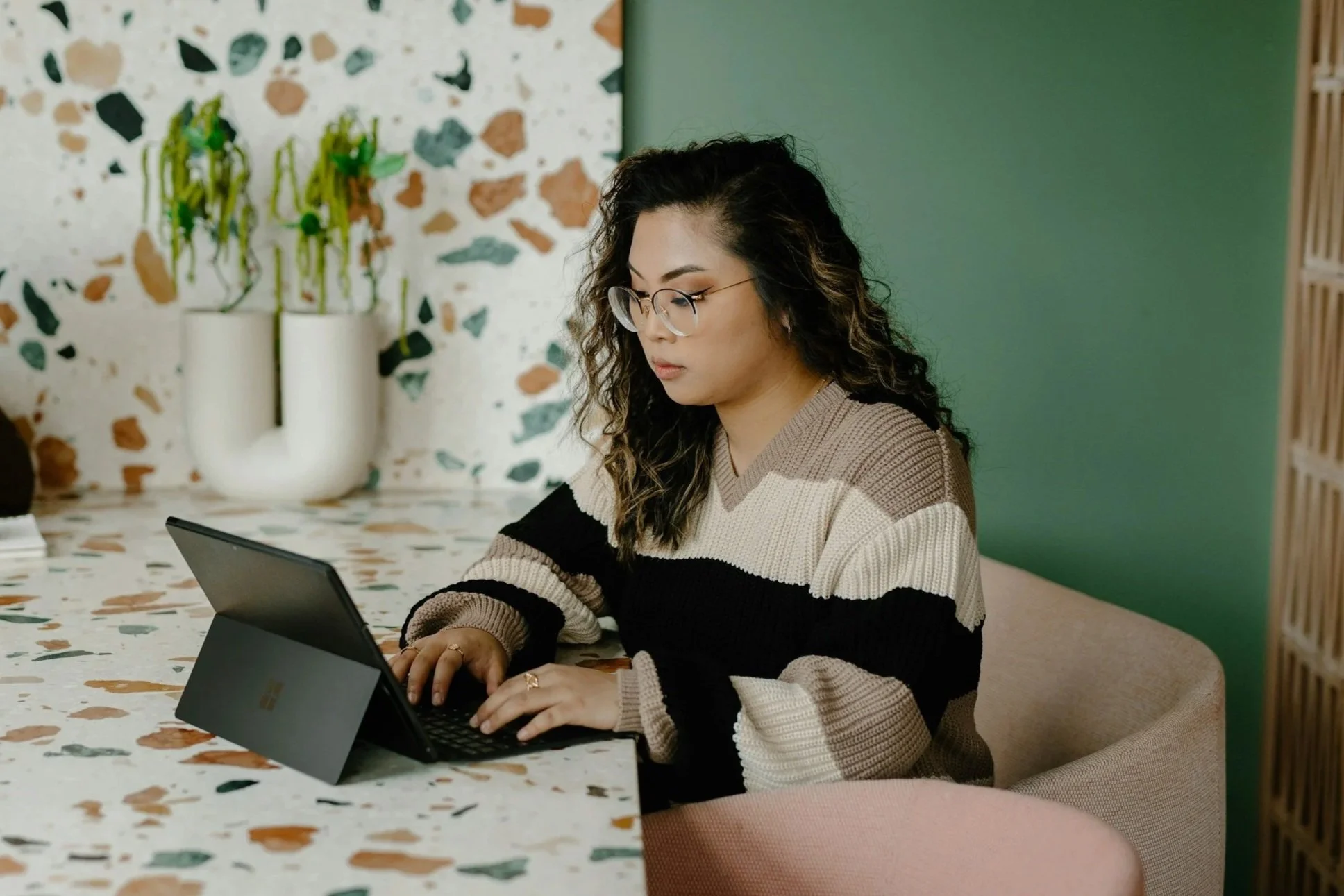A woman with curly hair and glasses sitting at a terrazzo table, using a tablet. In the background, there are two white vases with hanging plants and a decorated wall. The setting appears to be a modern indoor space.