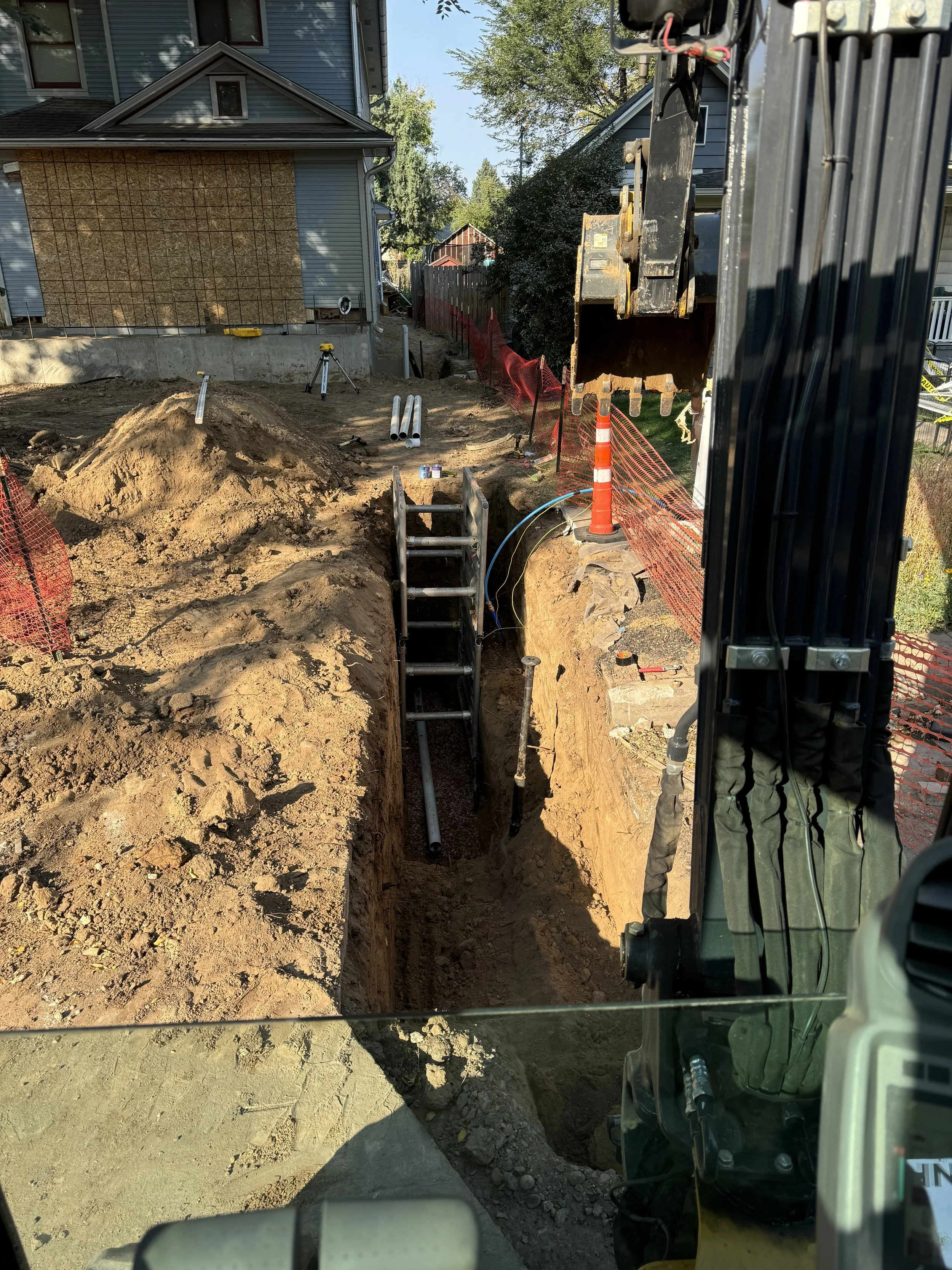 Construction site with a trench being dug, construction pipes, safety equipment, and a excavator arm visible, with a house and trees in the background.