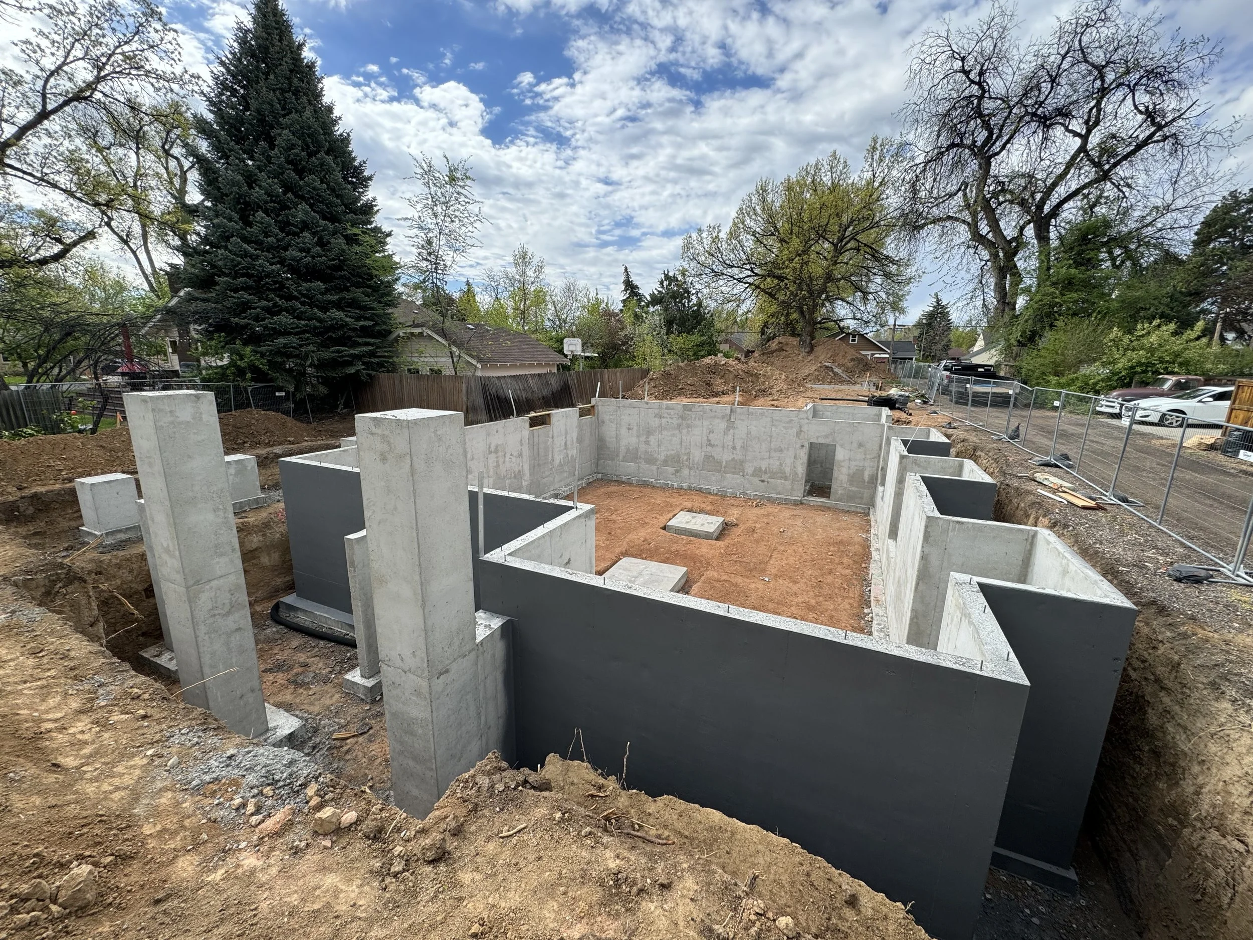 Construction site with concrete foundation walls and dirt, surrounded by fencing, with trees and houses in the background under a partly cloudy sky.