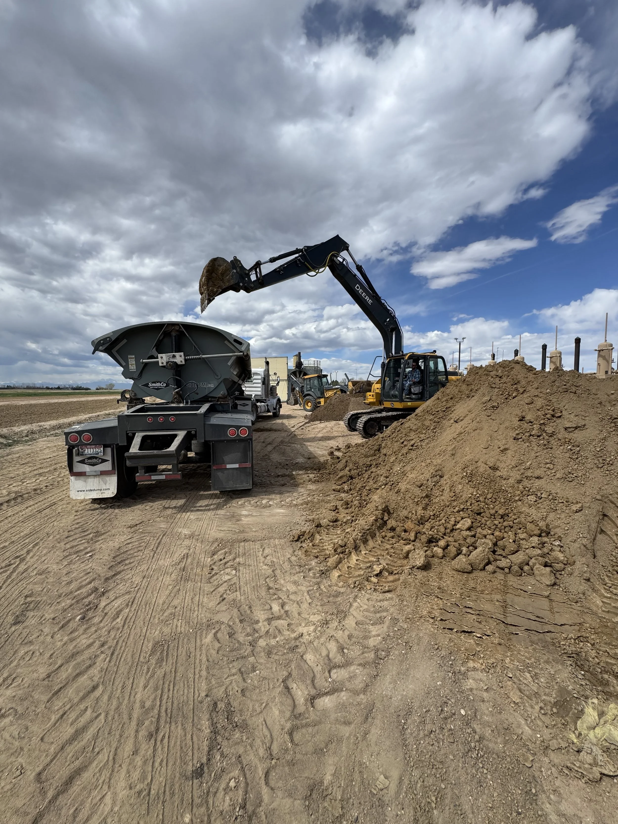 Construction site with a small excavator moving dirt and a flatbed truck nearby, under a partly cloudy sky.