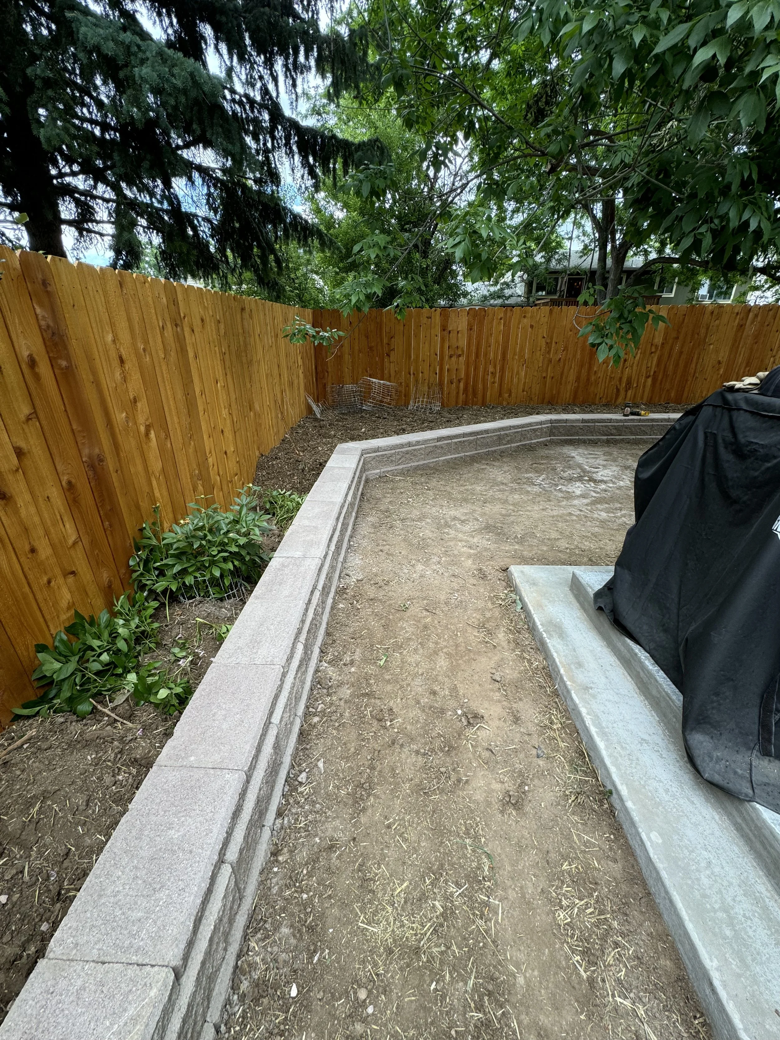 A backyard with a new wooden fence, some plants, and a stone border. Dirt area with some construction tools nearby.
