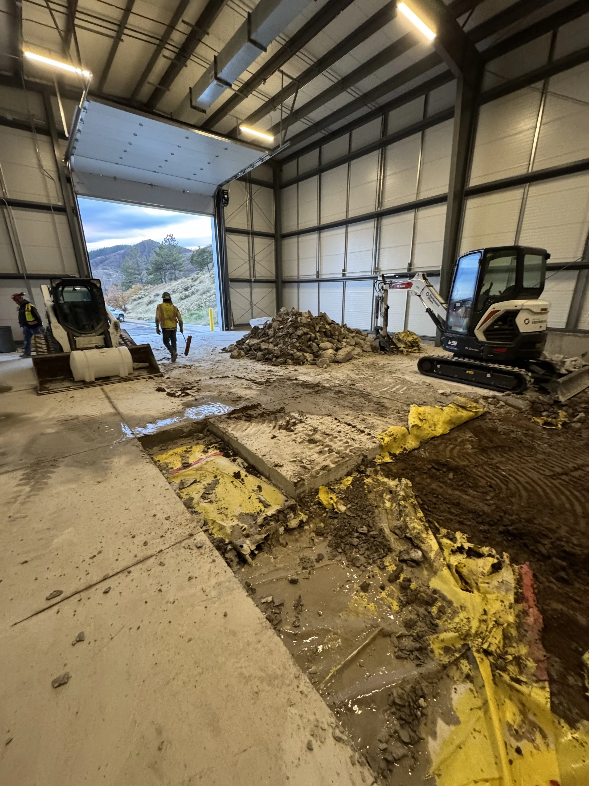 Construction workers inside a large industrial building with a partially open garage door, dirt and debris on the floor, a small excavator, and a pile of rocks.