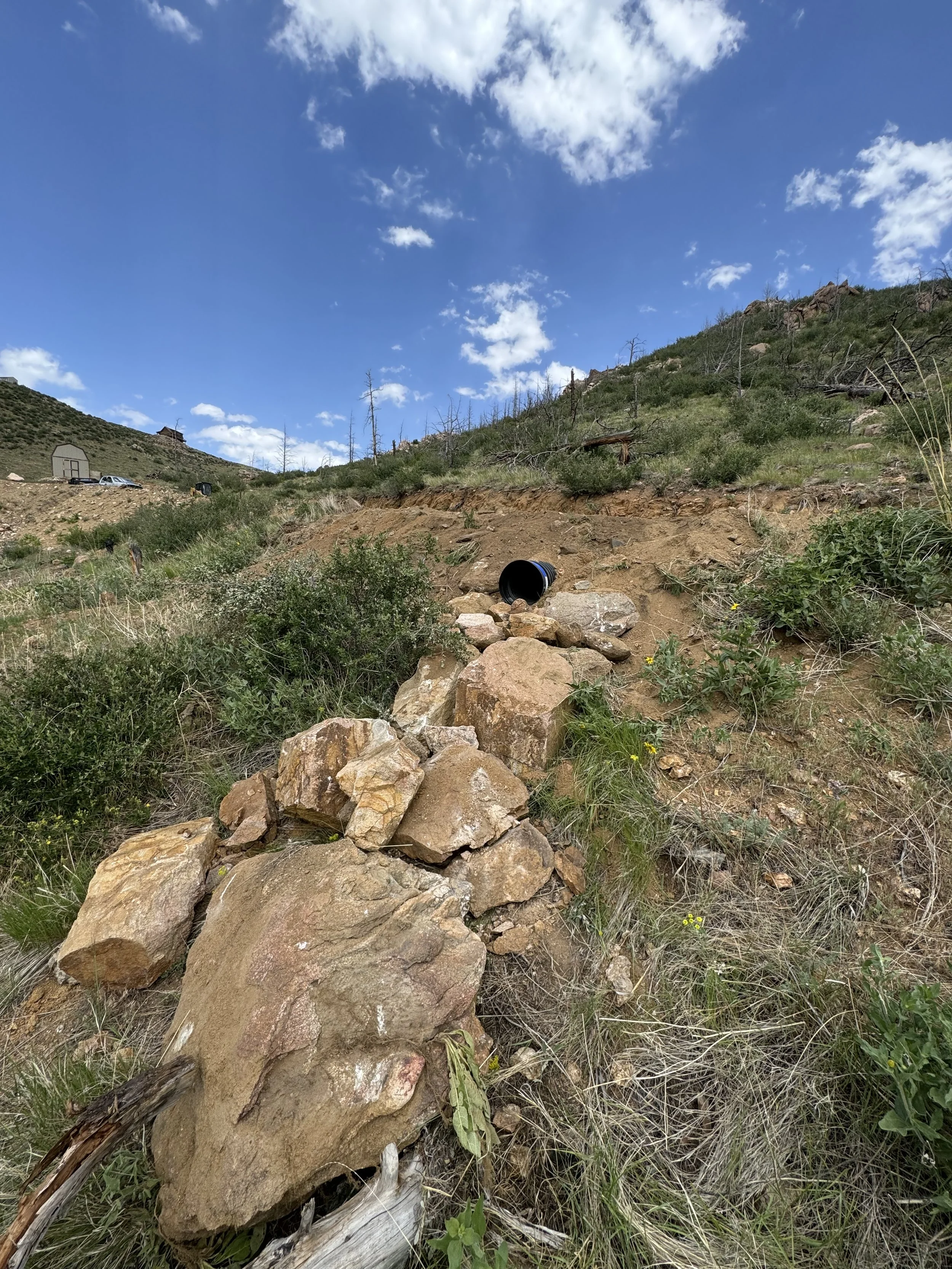 A hillside with rocks, some dry grass, green bushes, and a black pipe protruding from the ground in the foreground. The sky above is partly cloudy with a few scattered white clouds.