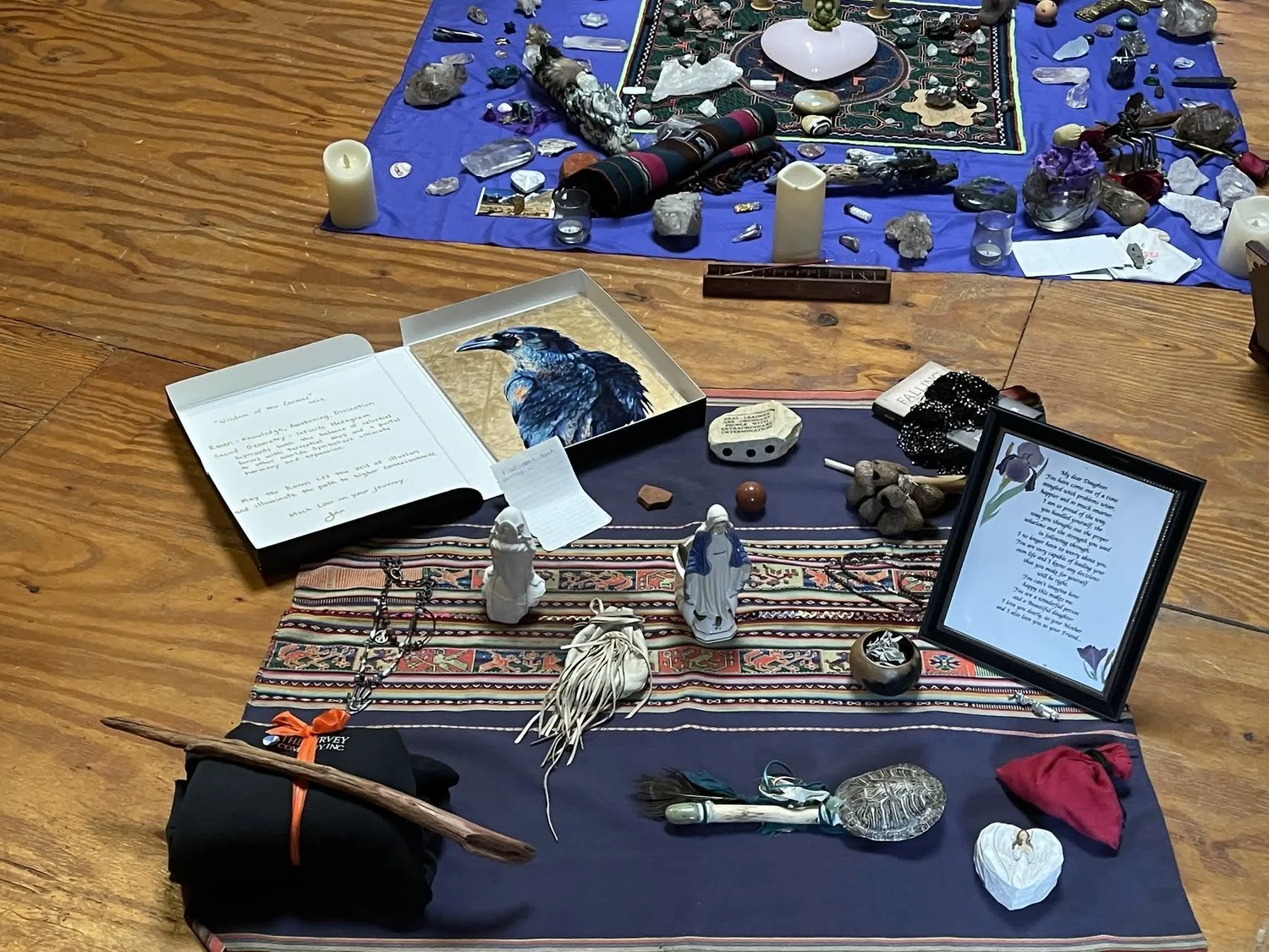 A spiritual altar setup on a wooden floor containing candles, crystals, a framed poem, a painted bird image, small statues, a decorative cloth, and various symbolic objects.