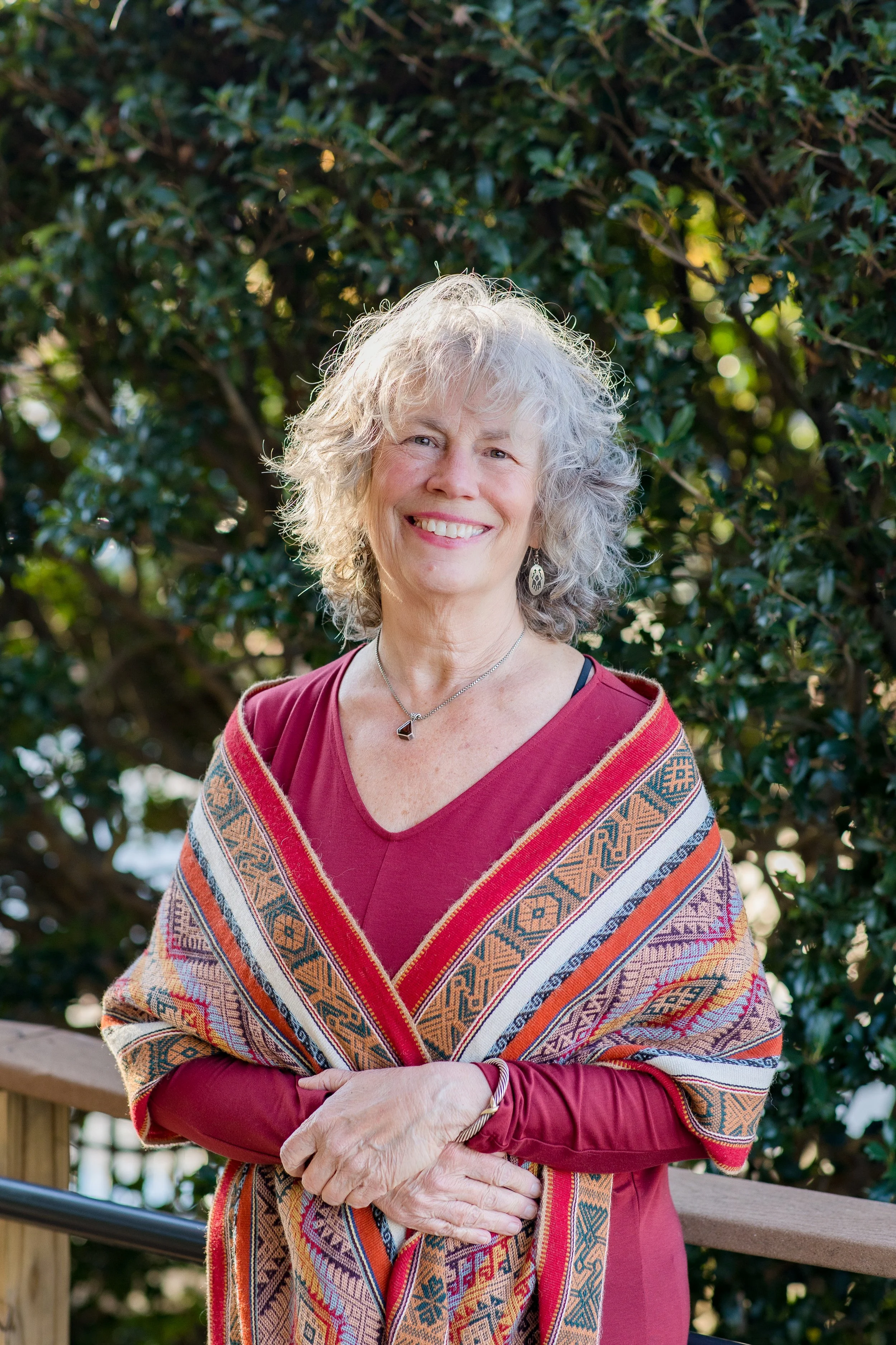 Older woman with gray curly hair, wearing a red top and colorful patterned shawl, standing outdoors in front of green foliage and trees.