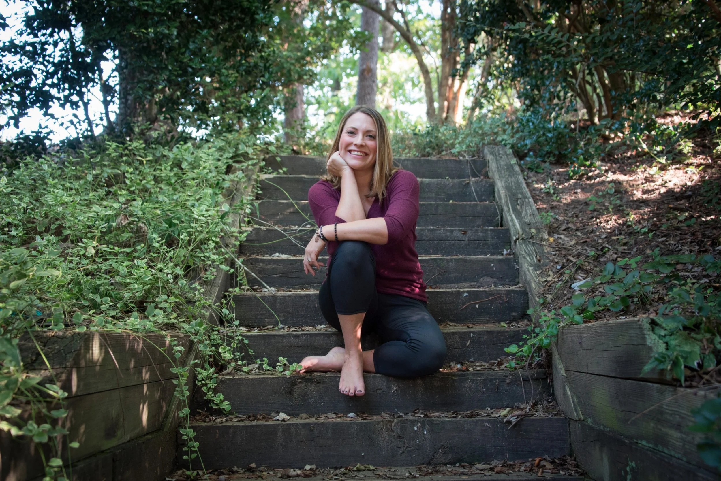 Woman sitting on outdoor wooden stairs surrounded by greenery, smiling with her chin resting on her hand.