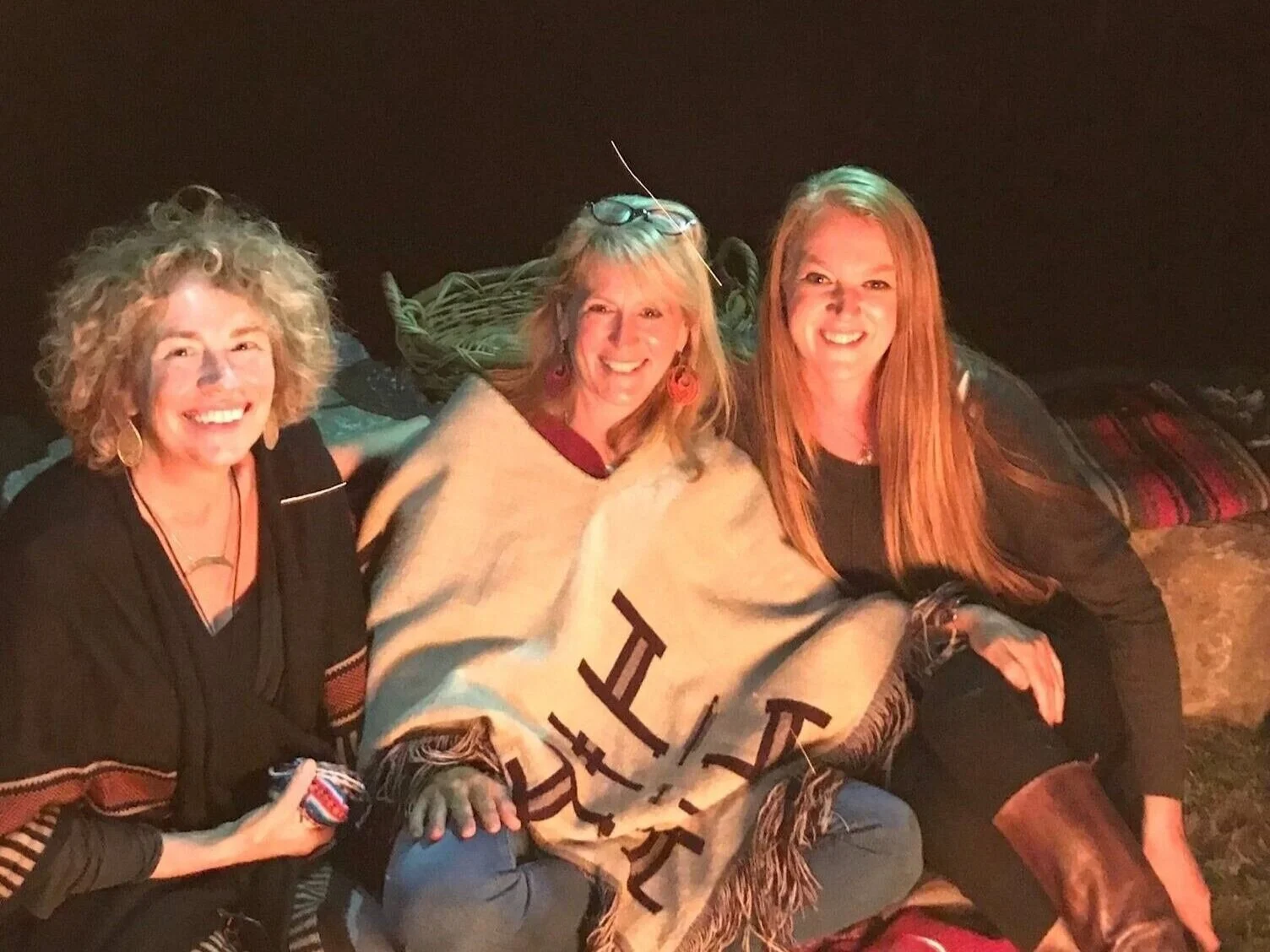 Three women sitting by a campfire, smiling, wrapped in blankets, with warm lighting and a dark background.