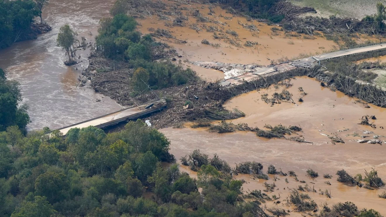 Flooded landscape with a partially collapsed bridge and debris in muddy water, surrounded by trees.