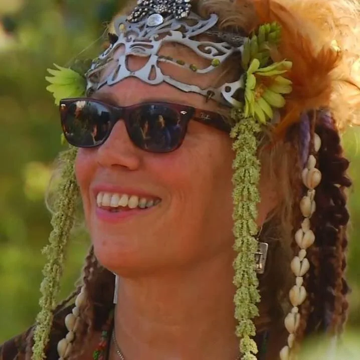 Person wearing sunglasses and a decorative headpiece with flowers and shells, smiling outdoors.
