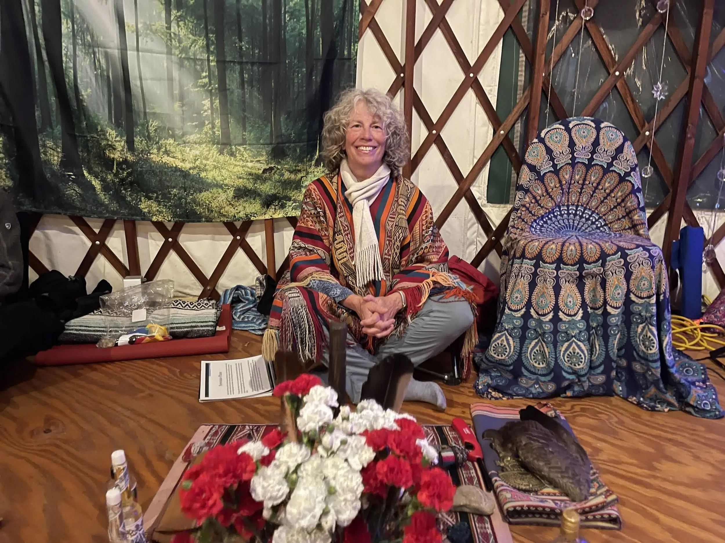 Smiling person sitting in a decorated yurt, surrounded by colorful fabrics and items including a chair, flowers, and a backdrop of a forest scene.