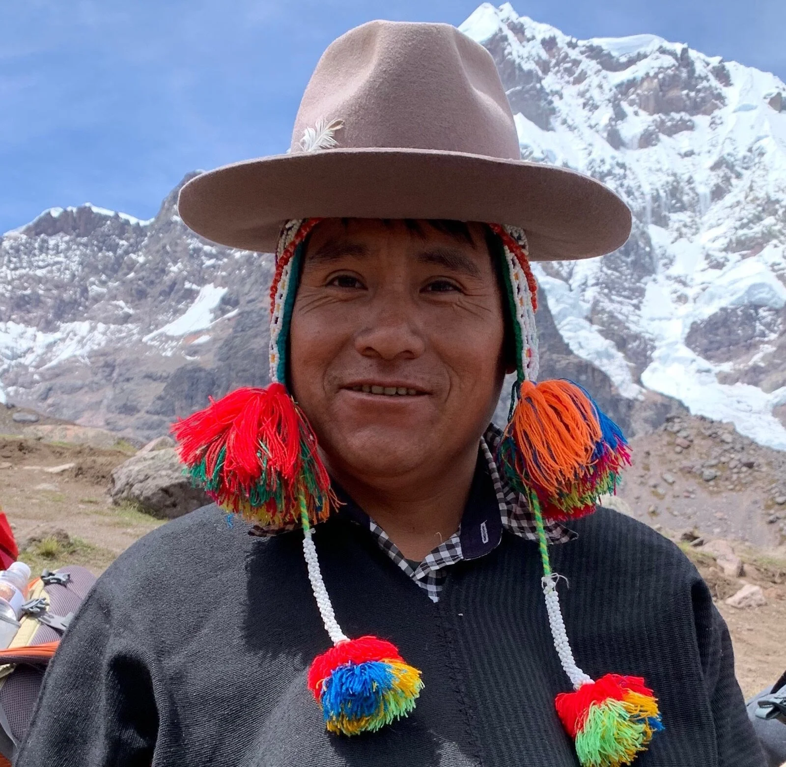 Person wearing a traditional hat with colorful tassels, standing in front of snow-covered mountains.