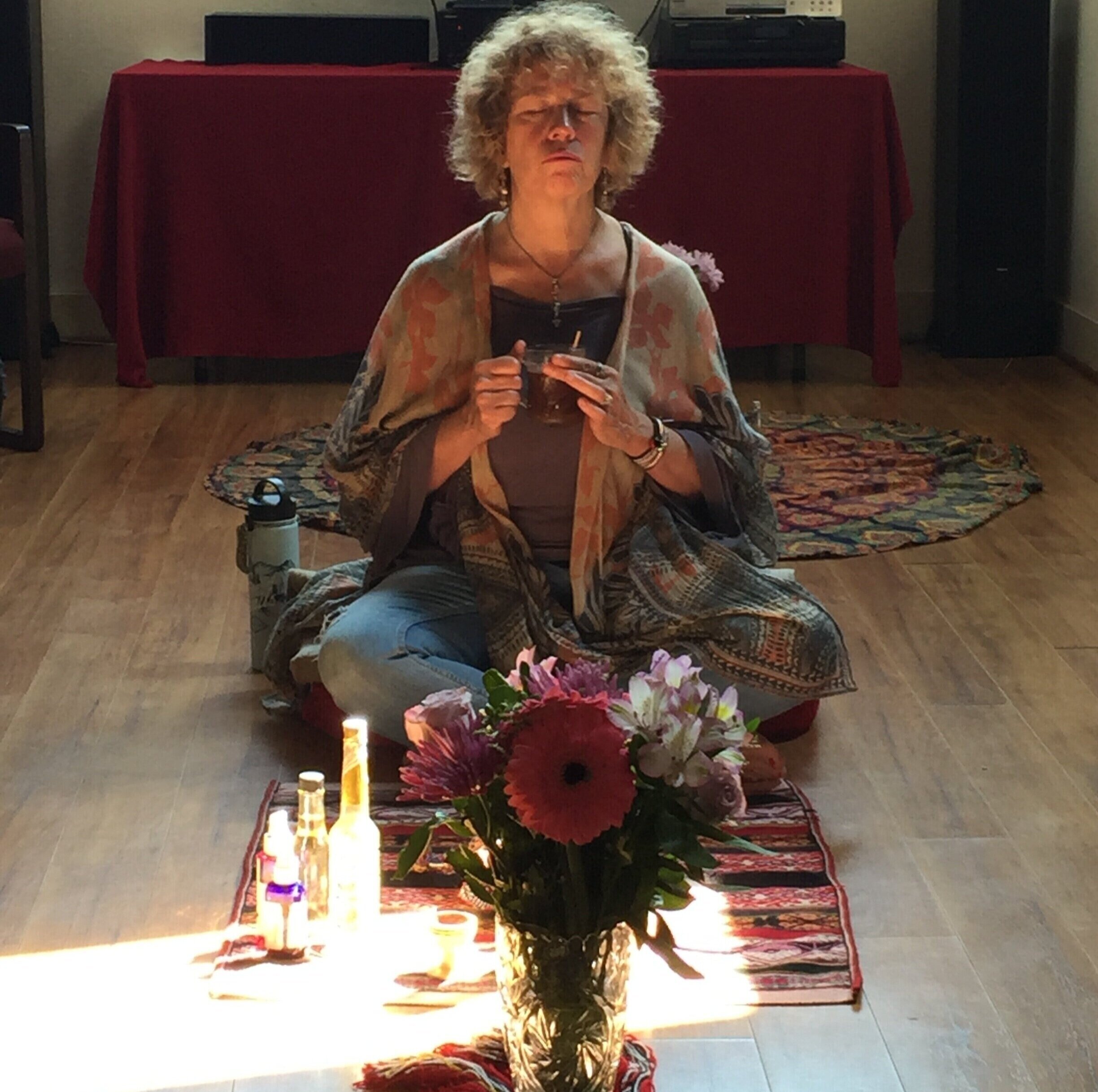 A person meditating indoors, sitting cross-legged on a mat. They are holding a small vessel and have their eyes closed. In front of them is a small arrangement with flowers and bottles. The room has wooden flooring and a red tablecloth in the background.