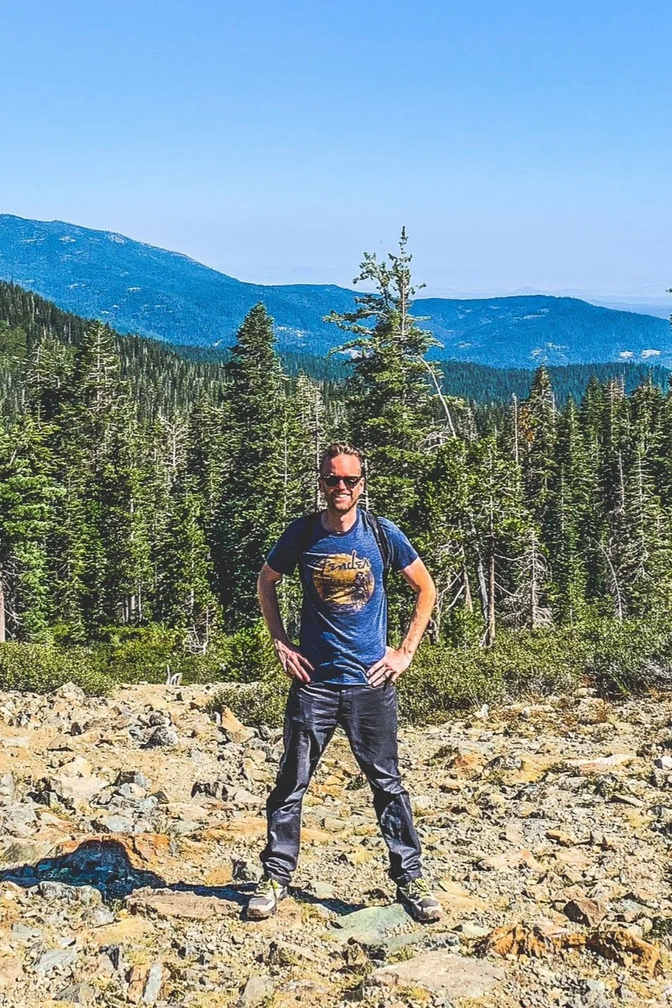 Man in a blue shirt and sunglasses standing in a rocky landscape with a forest and mountains in the background.