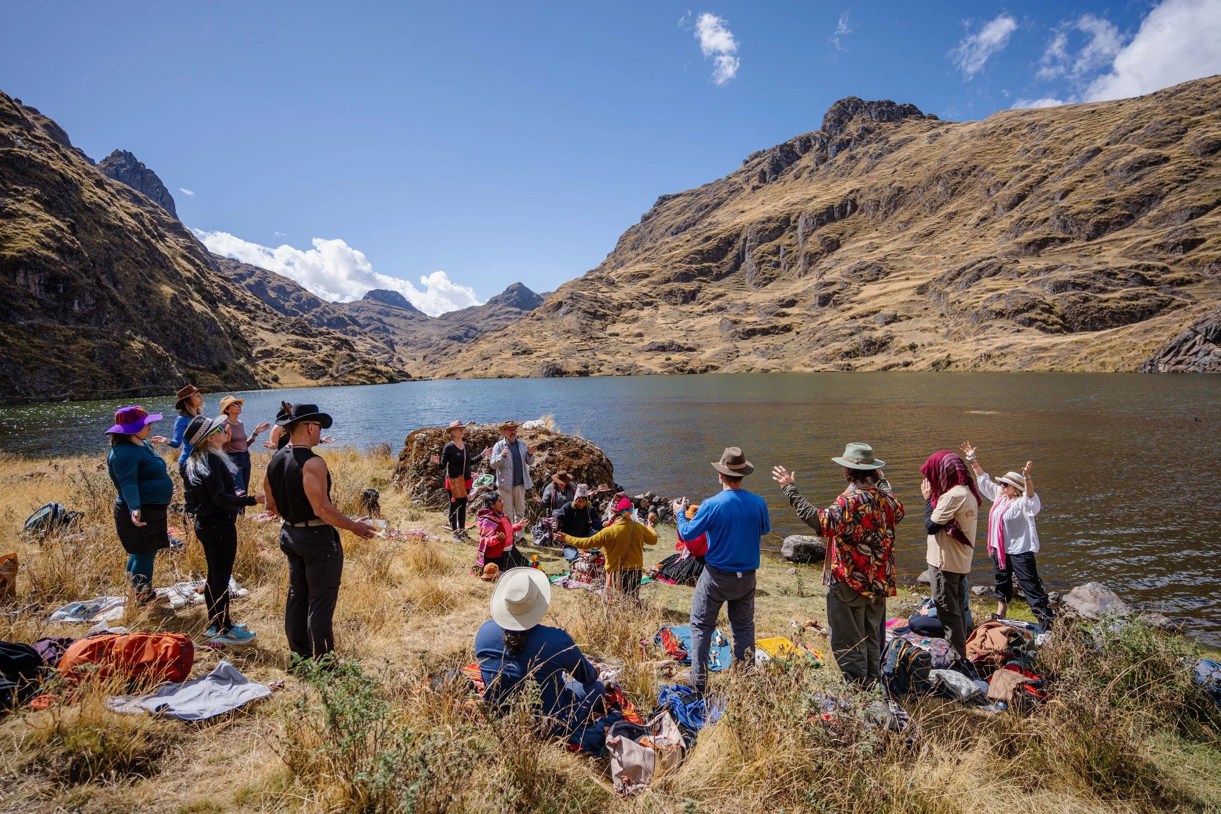 Group of people standing by a lake in a mountainous area, participating in an outdoor gathering or ritual, with clear blue skies.