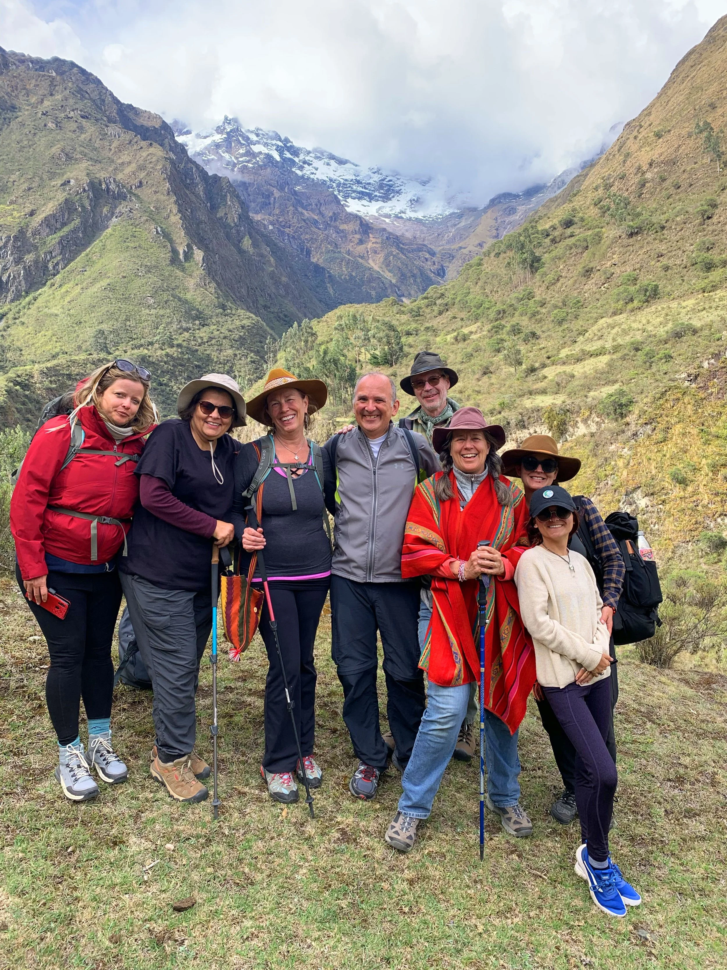 Group of hikers posing in front of mountain landscape wearing casual outdoor clothing and hats, under cloudy sky with snow-capped peaks in the background.