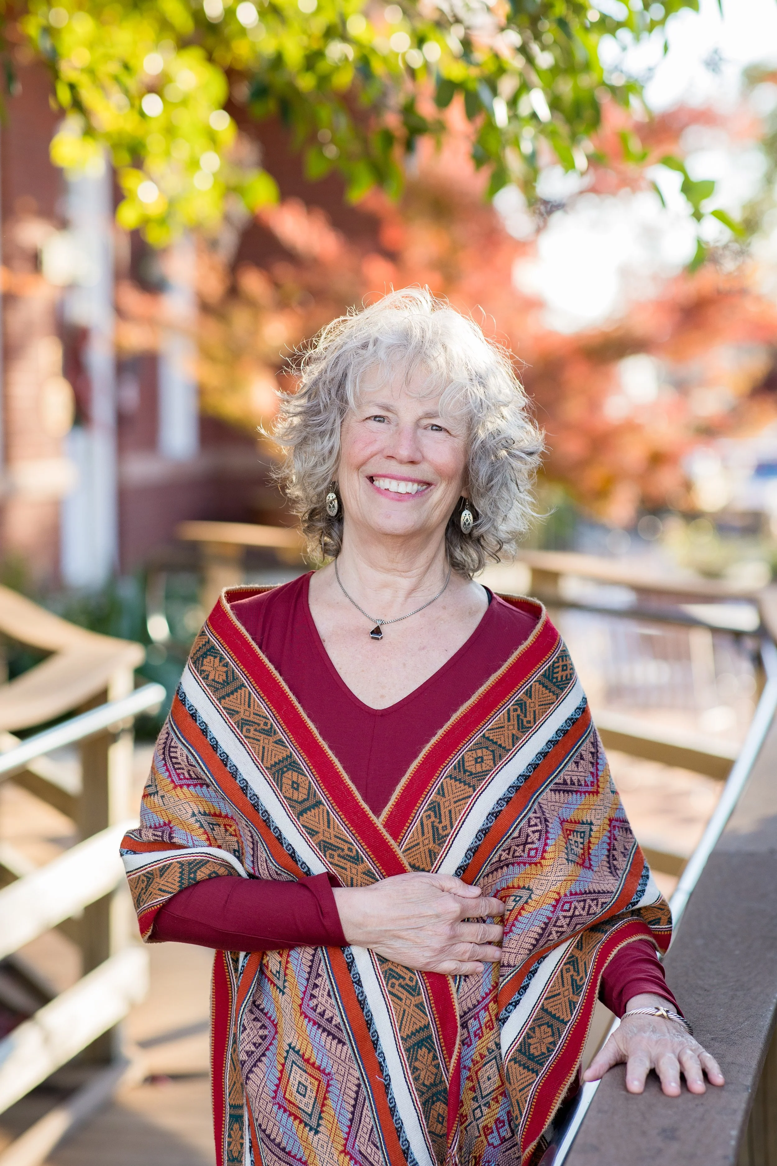 Smiling woman with gray curly hair wearing a colorful shawl, standing outdoors with autumn leaves and a wooden railing in the background.