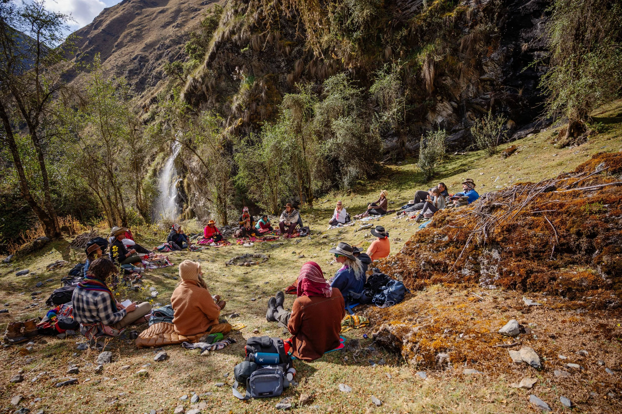 A group of people sitting outdoors on a grassy area surrounded by rocks, moss, and trees, with a waterfall visible in the background. They appear to be in a circle, possibly in a meeting or group discussion. They are dressed in casual and colorful clothing, some wearing hats.