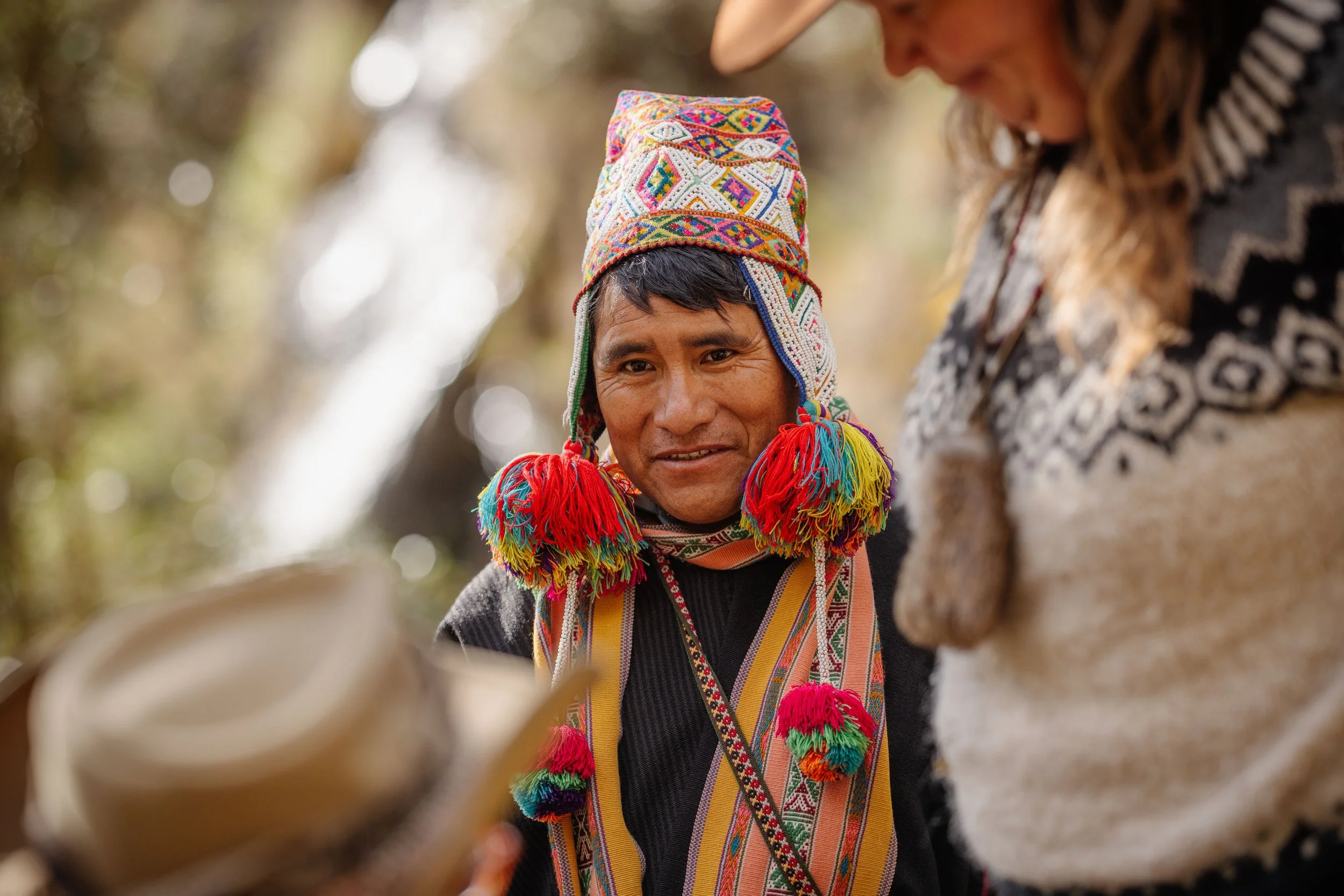 A person wearing traditional Andean clothing, including a colorful knitted hat with tassels, smiling outdoors. Another individual, partially visible, is nearby. The background shows blurred greenery.