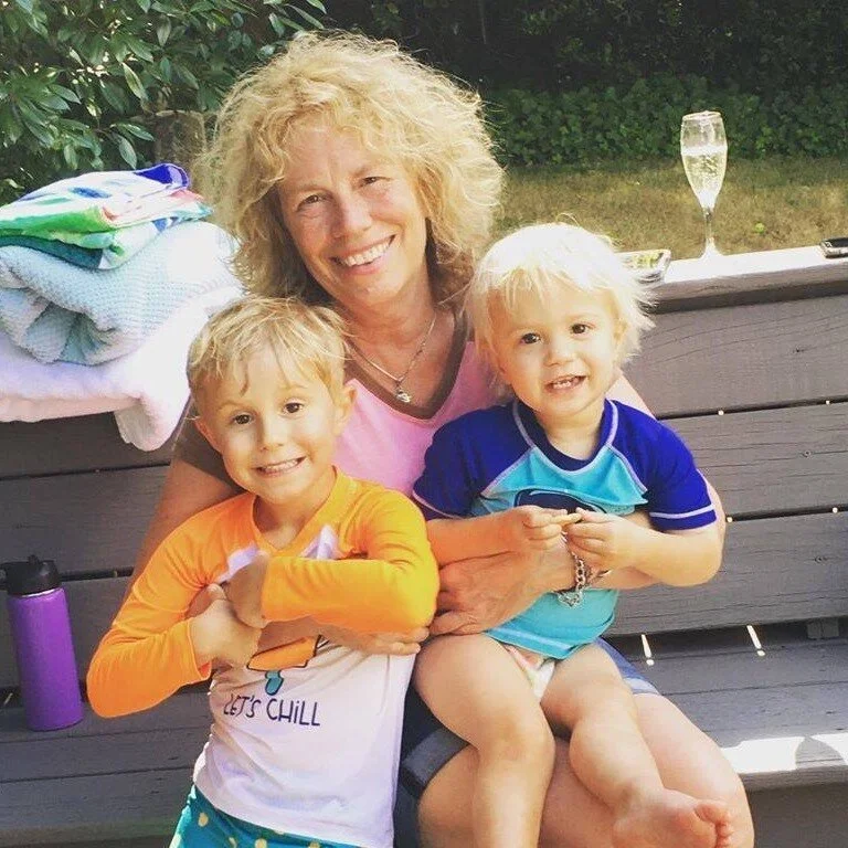 A smiling woman sits on a bench outdoors, hugging two young children. One child wears an orange and white rash guard, the other a blue one. A stack of towels and a glass of champagne are visible in the background.