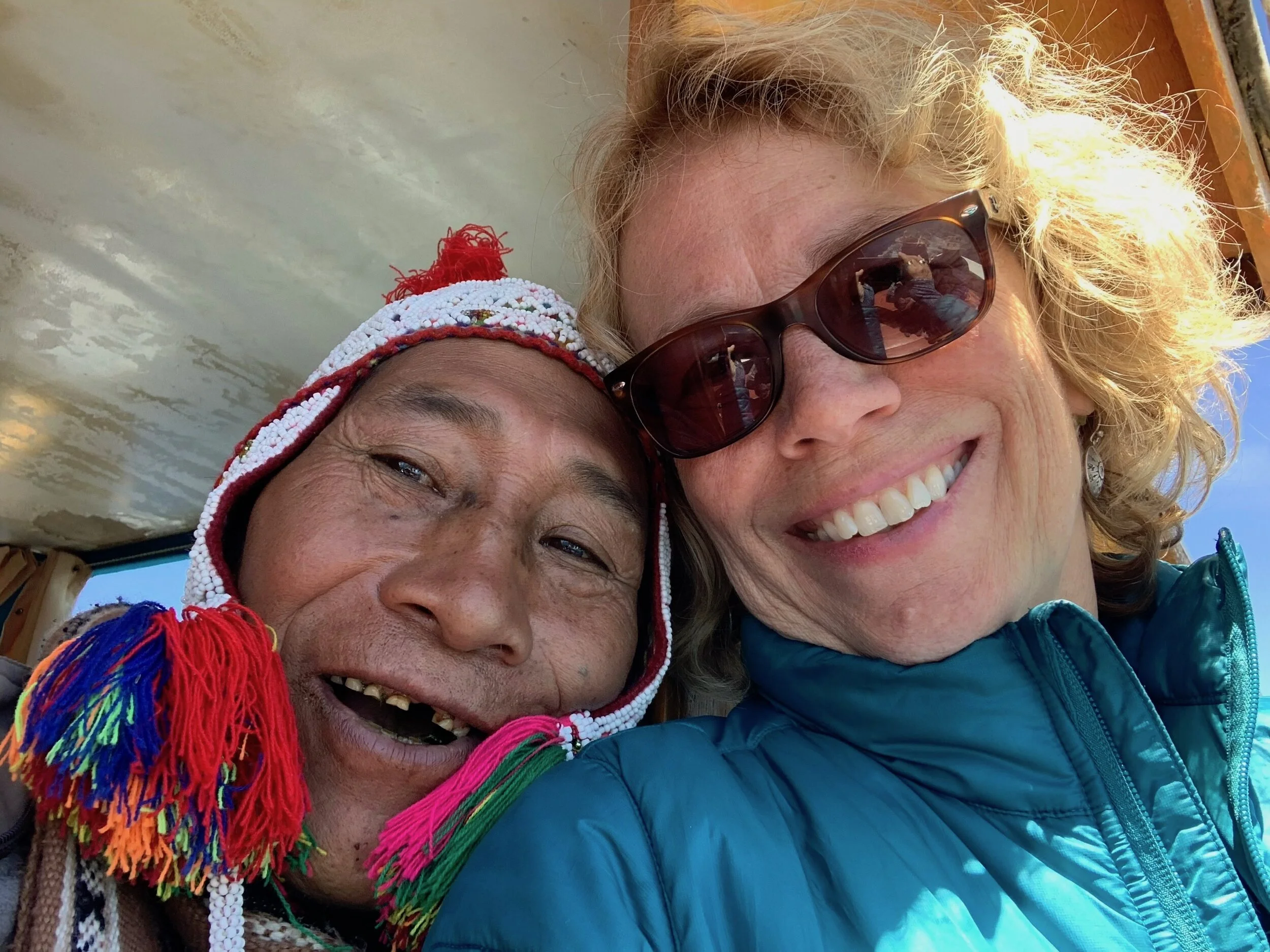 Two people smiling, one wearing a colorful traditional hat, the other with sunglasses and a teal jacket.