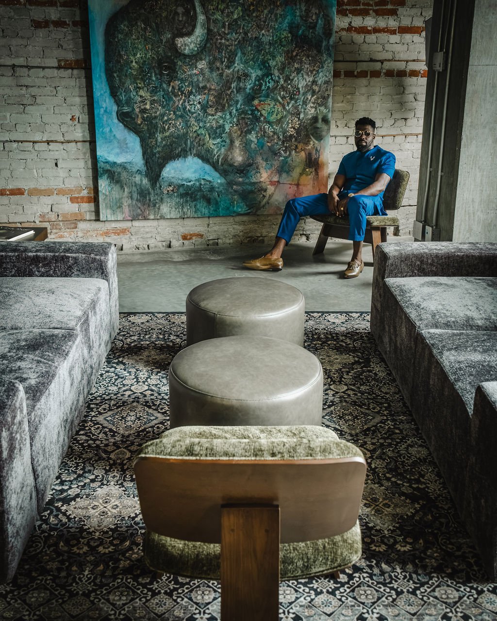 A man in a blue outfit and sunglasses sits on a wooden chair with a patterned cushion, in an industrial-style room with a brick wall. Behind him is a colorful large painting of a buffalo's head, and in the foreground are plush seating and ottomans on a decorative rug.