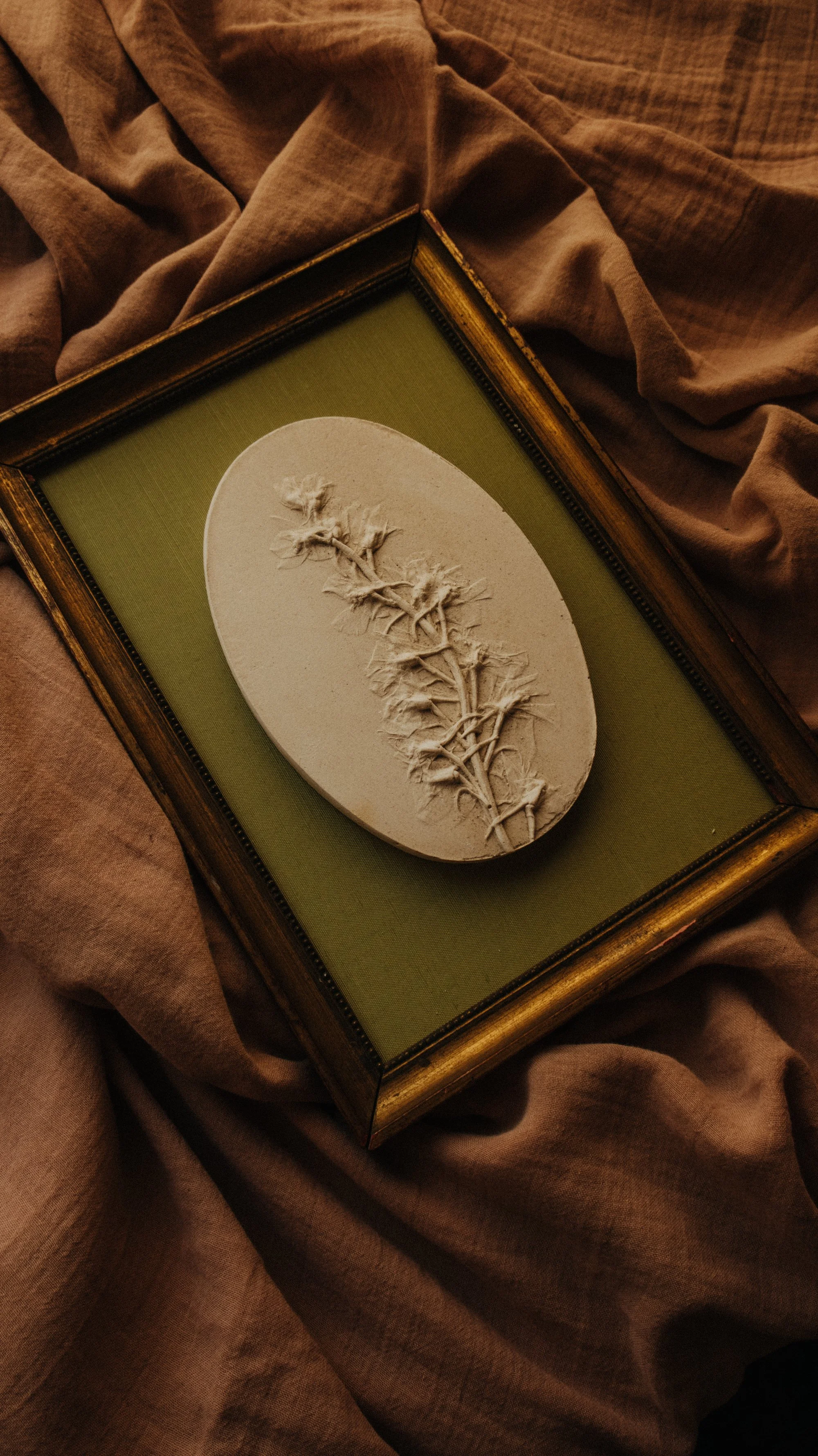 Oval plaster bas-relief of a single larkspur stem mounted in a vintage gilt frame with olive-green fabric.