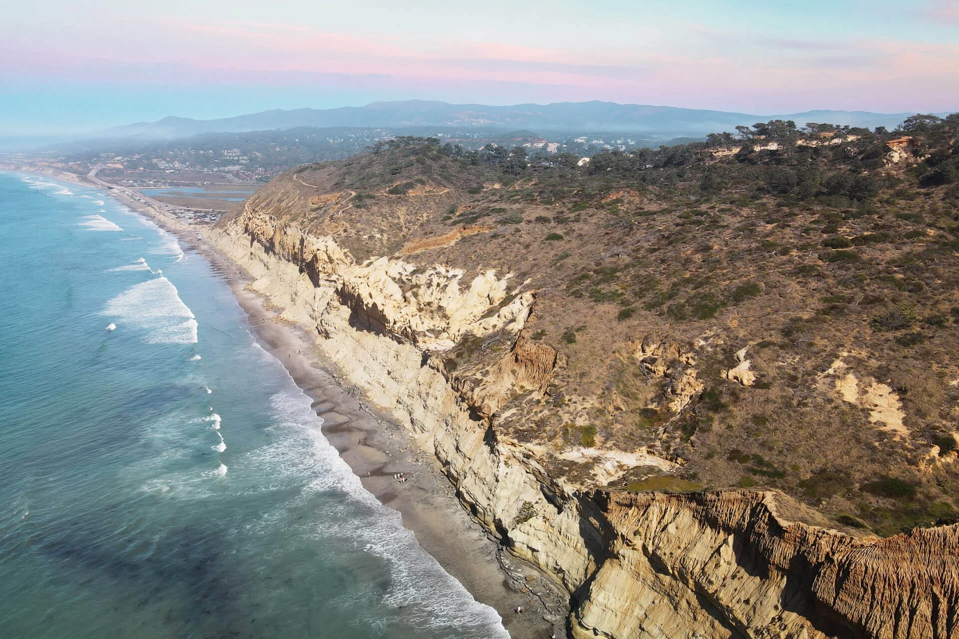 An aerial view of a rugged coastline with cliffs, a sandy beach, and the ocean, with mountains in the background during sunset.