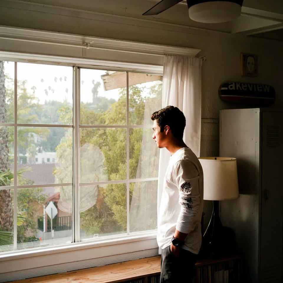A man standing by a window in a sunlit room, looking out thoughtfully. The setting is calm and grounded, reflecting the quiet space provided in men’s counselling sessions.