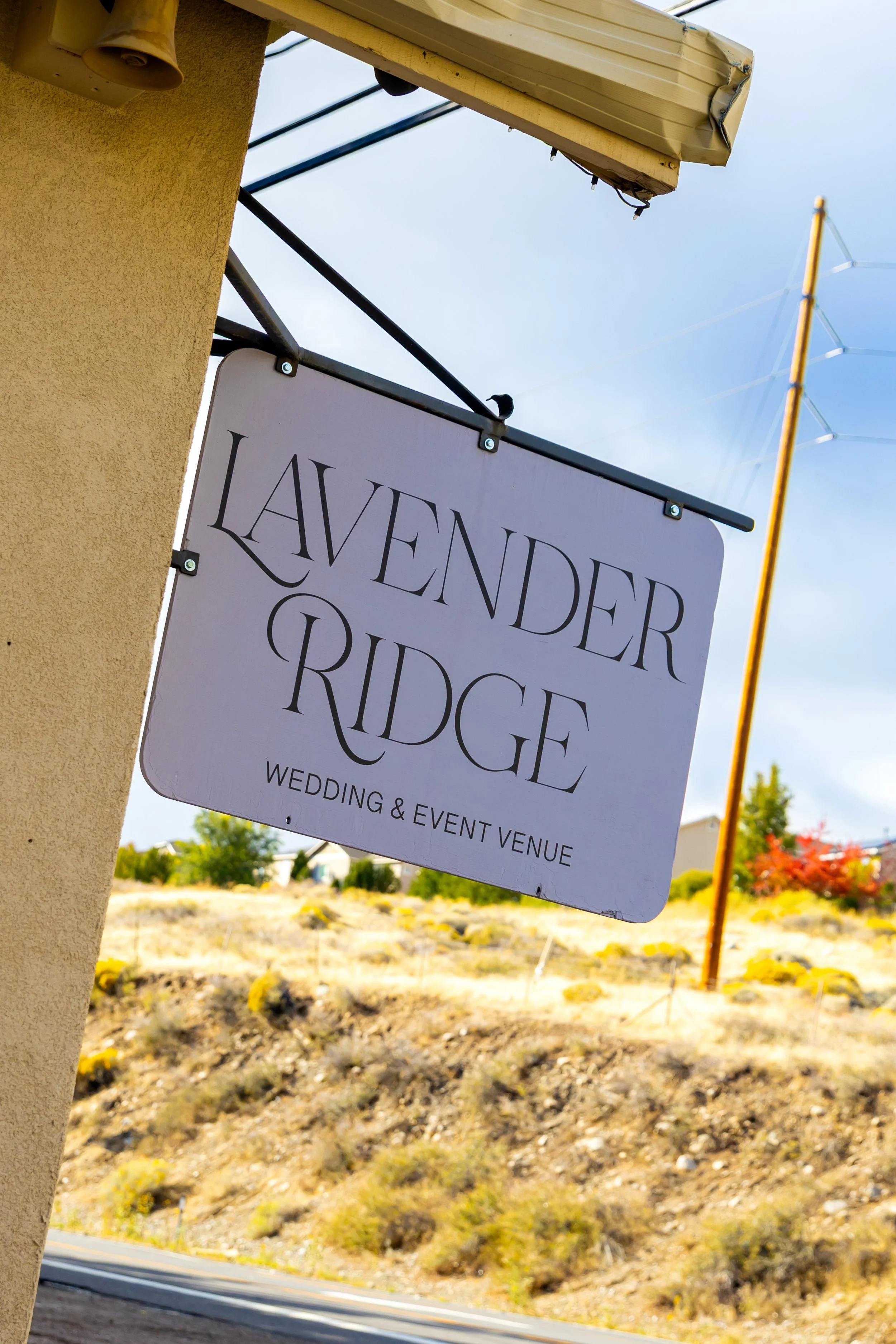 Sign for Lavender Ridge wedding and event venue hanging from a building, with a hillside and power line in the background.