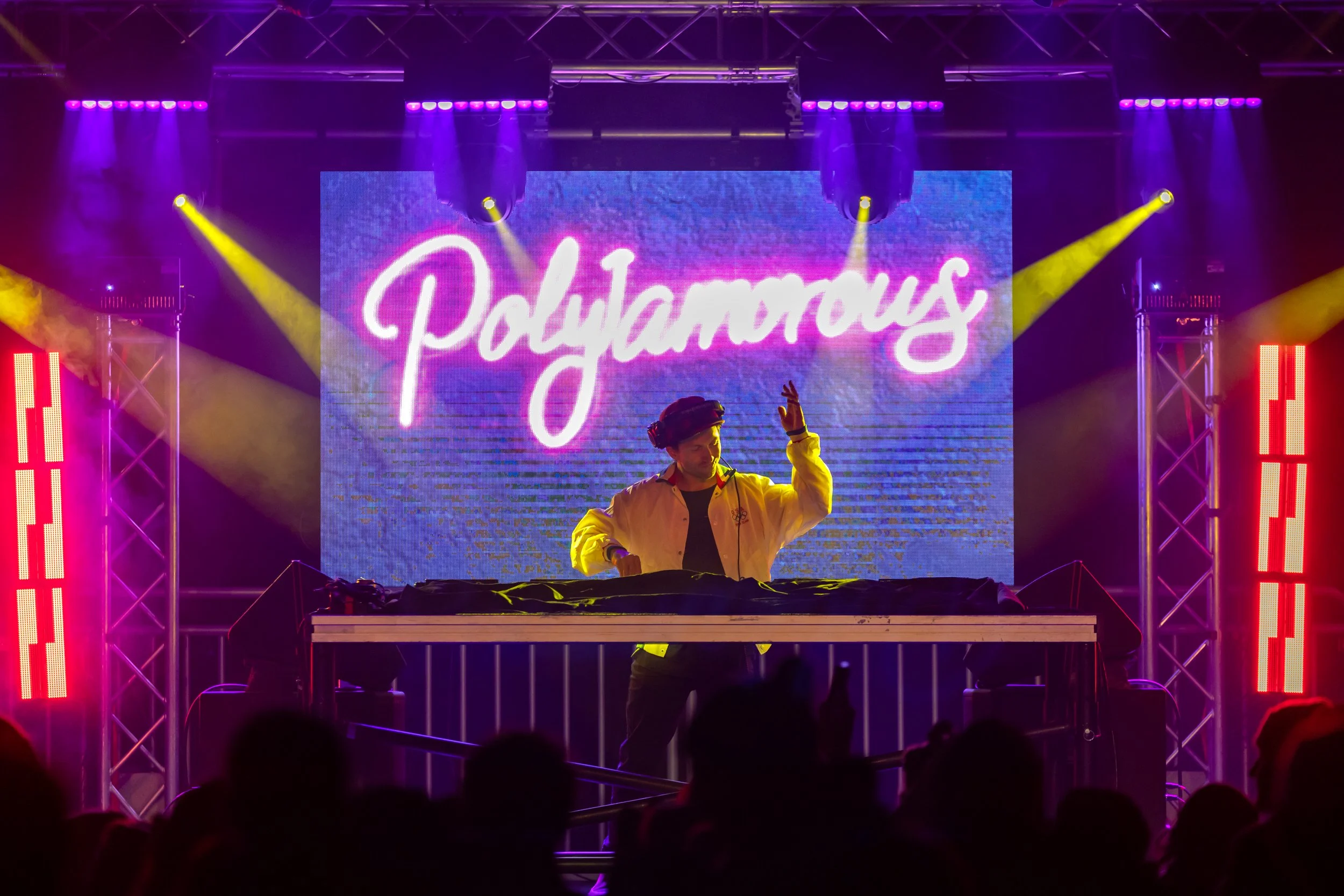 A DJ performs at a nightclub with vibrant purple and yellow stage lighting. Behind him is a large LED screen displaying the word 'Polyramus' in neon cursive font. The DJ wears a yellow jacket and a black cap, and his hand is raised in the air.