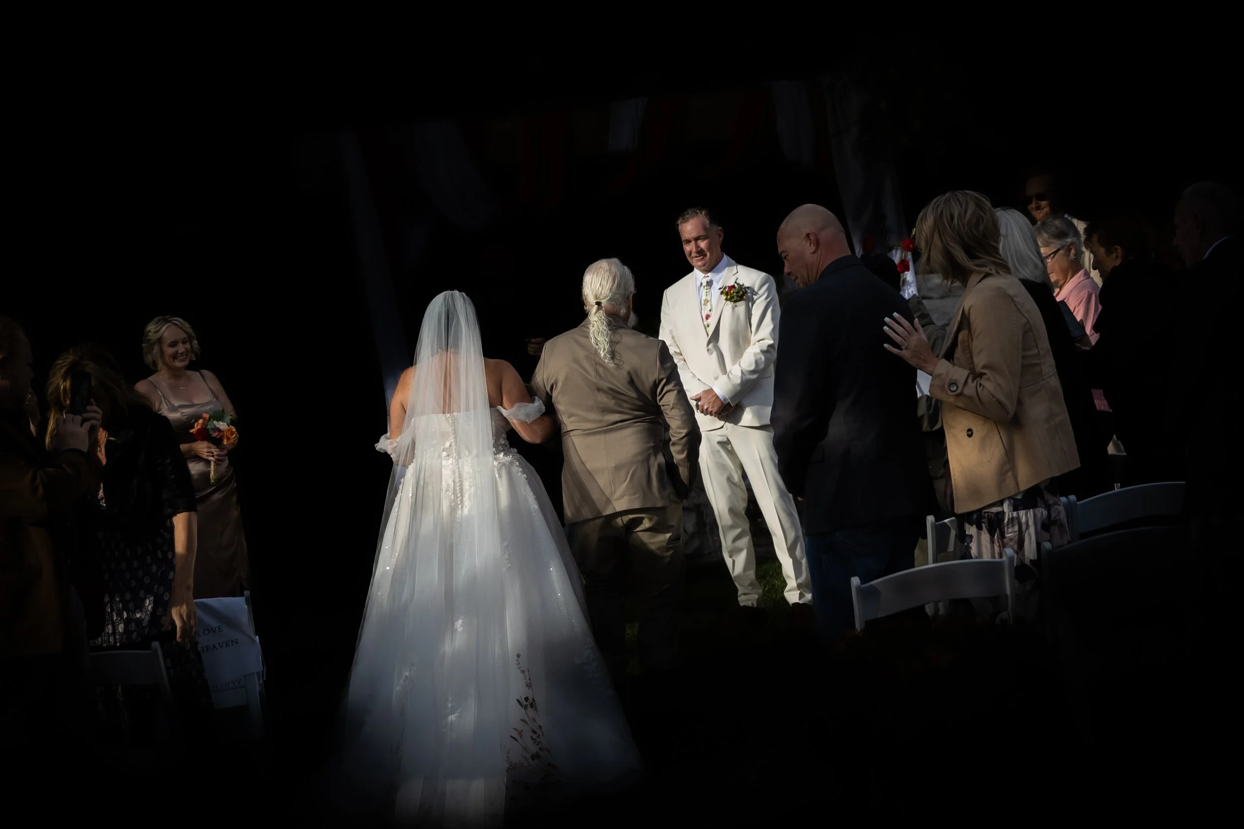 A wedding ceremony with the bride, in a white wedding gown and veil, and the groom, in a white suit, exchanging vows on a stage with guests standing around, some taking photos and others observing.