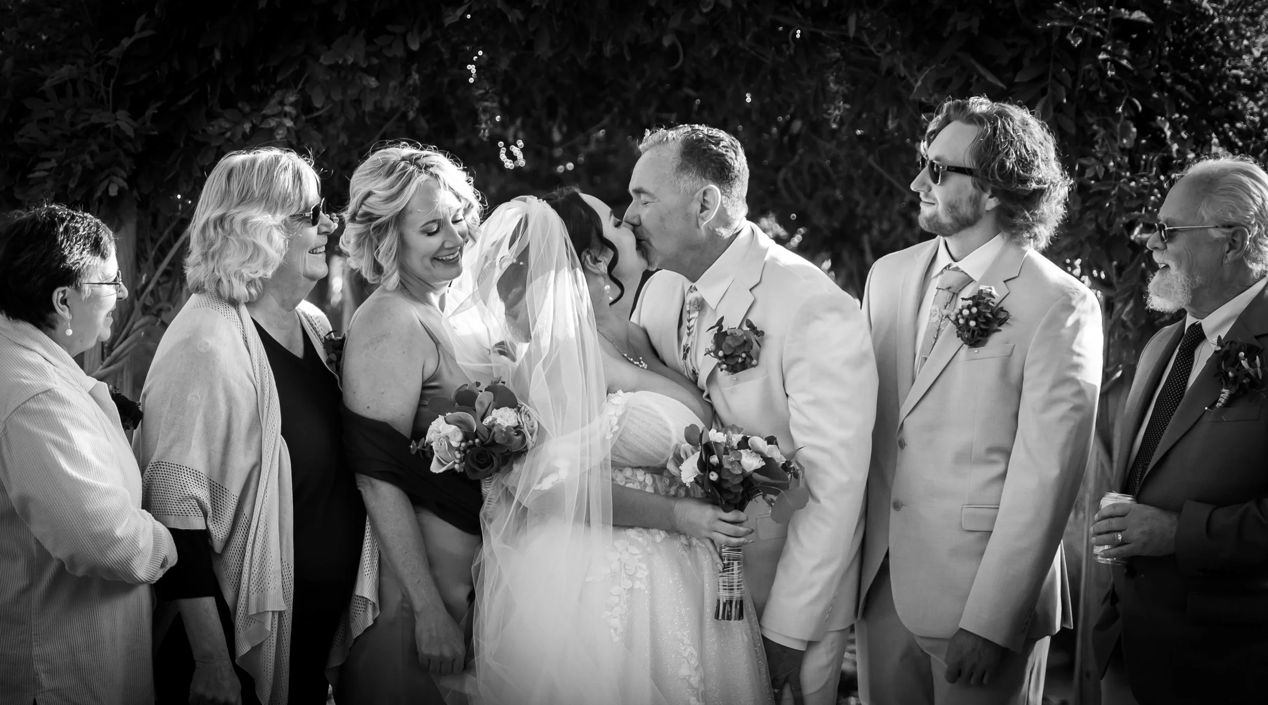 A black-and-white photograph of a wedding scene with a bride and groom sharing a kiss, surrounded by family and friends outdoors. The bride is wearing a wedding dress with a veil and holding a bouquet, while the groom is in a suit. The wedding party 